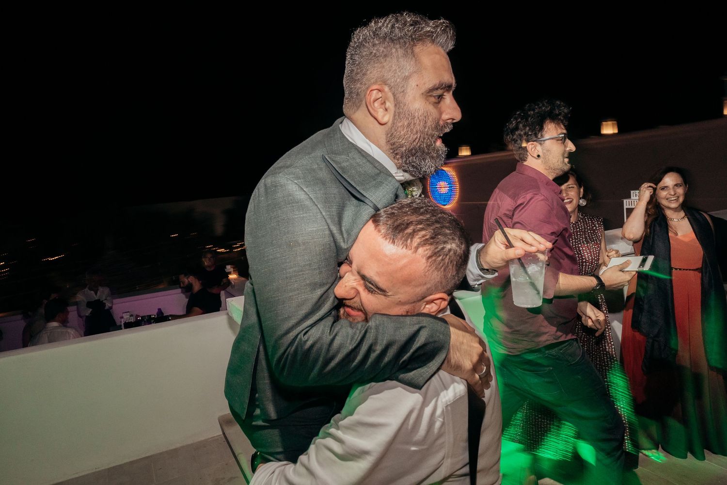 Party guests embracing and dancing during a lively wedding reception with green lighting.