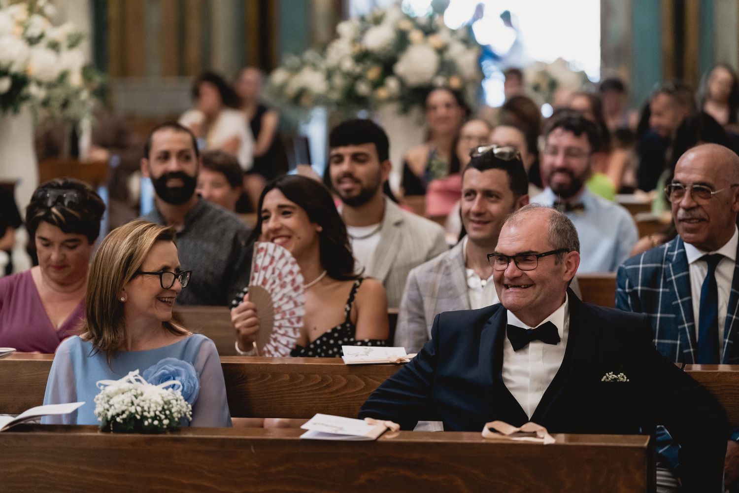 Wedding guests sitting in church pews smiling and laughing during ceremony.