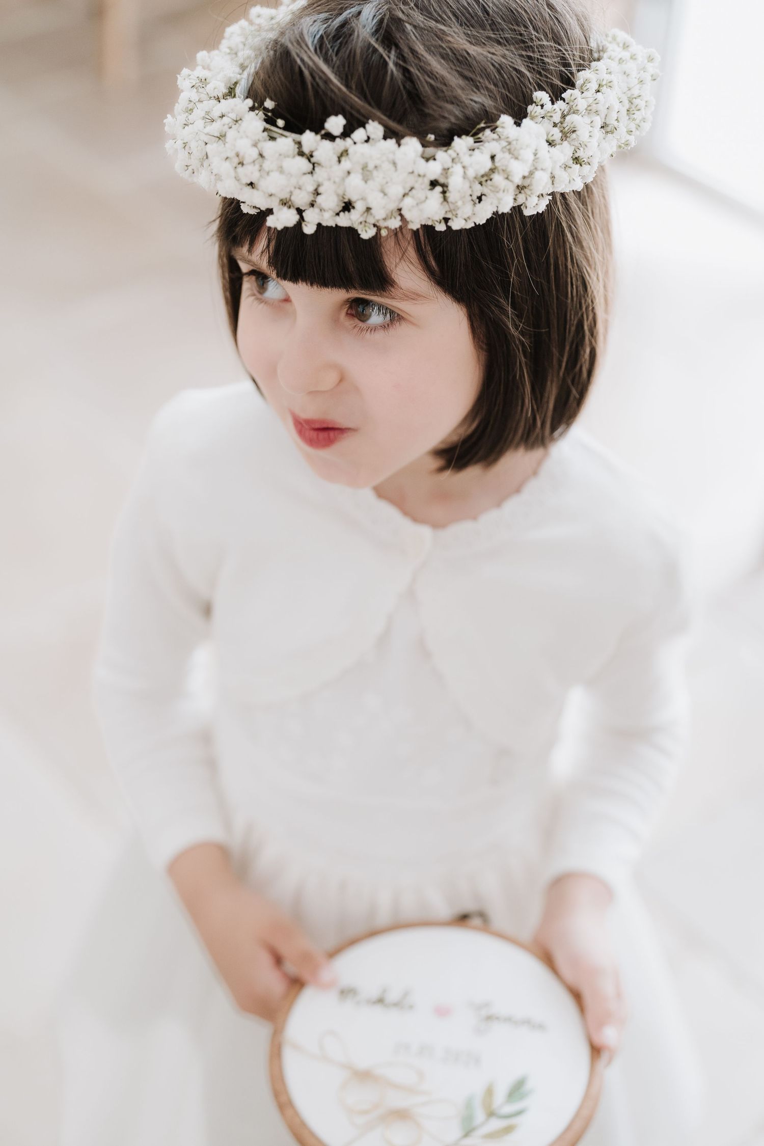 Child wearing white dress and floral crown holds an embroidered hoop.