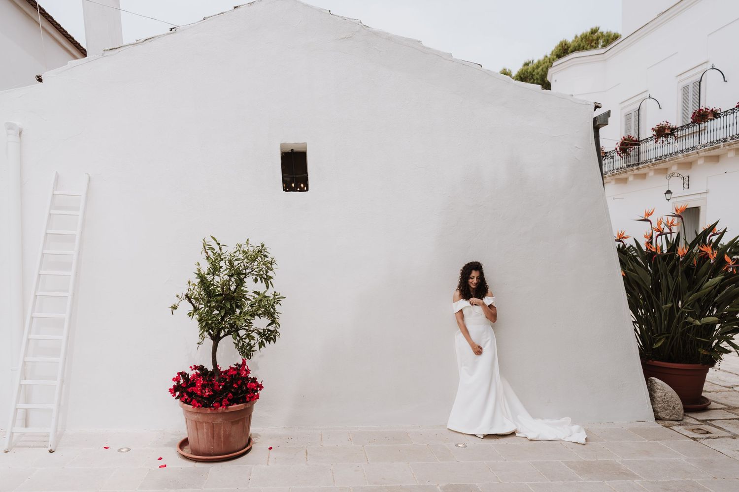 A bride in a stunning white dress stands alone against a minimalist white wall with potted plants.