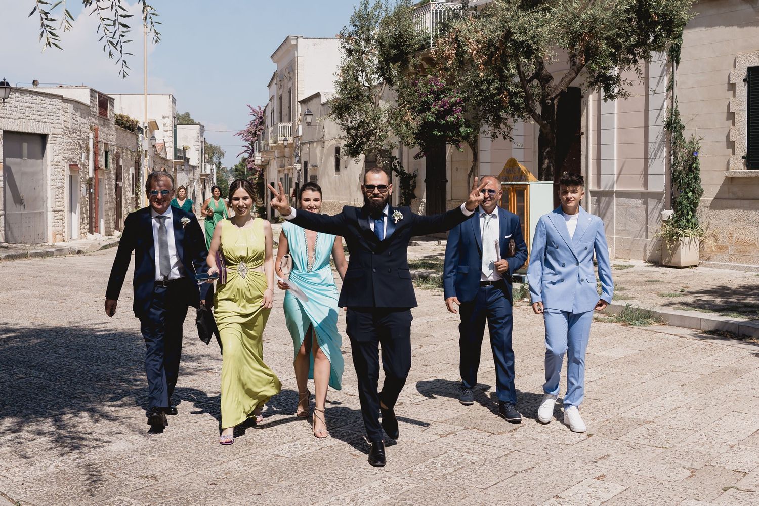 Wedding party walking down a sunny Mediterranean street in colorful attire.