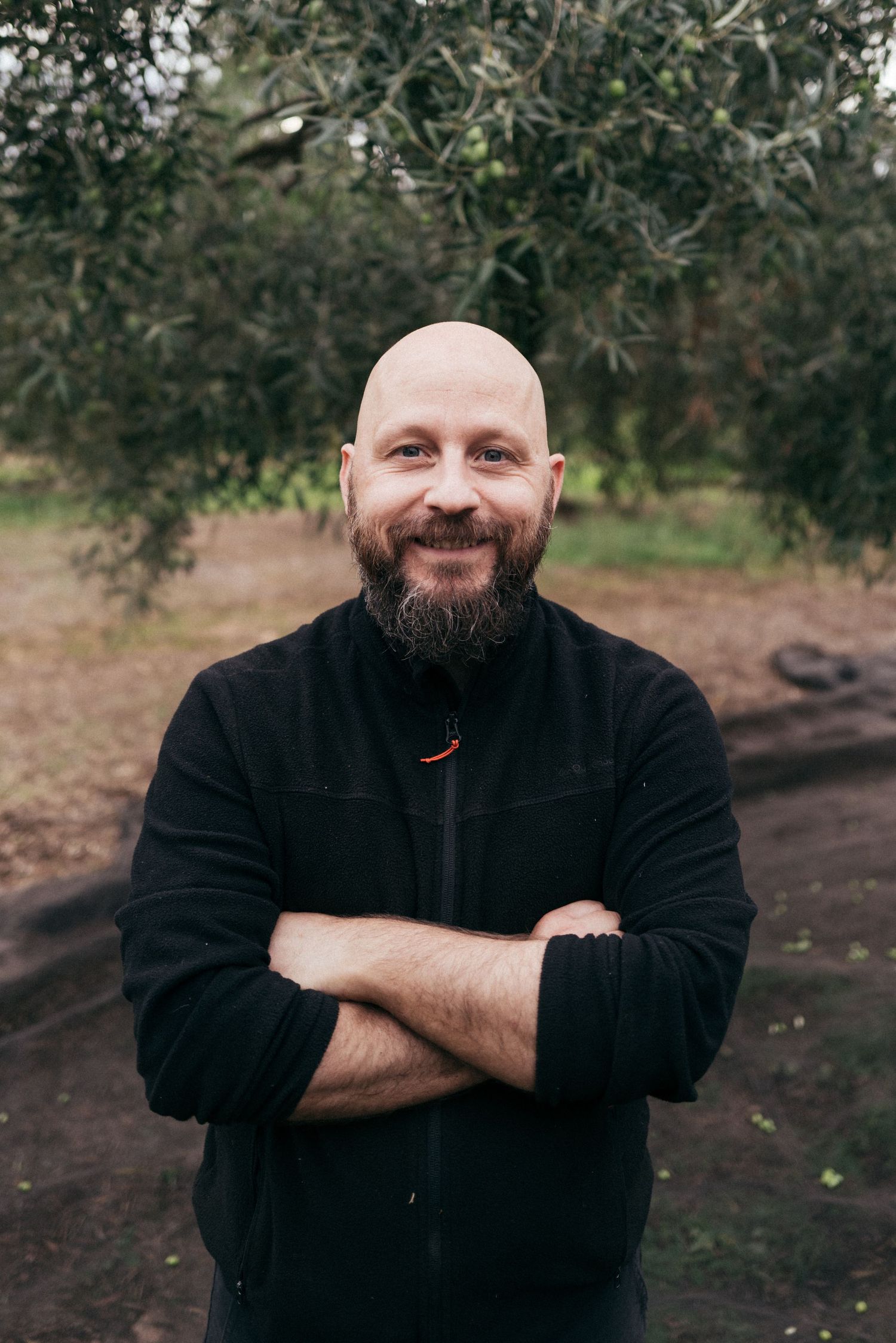 Person in black fleece jacket smiling with arms crossed against olive grove background.