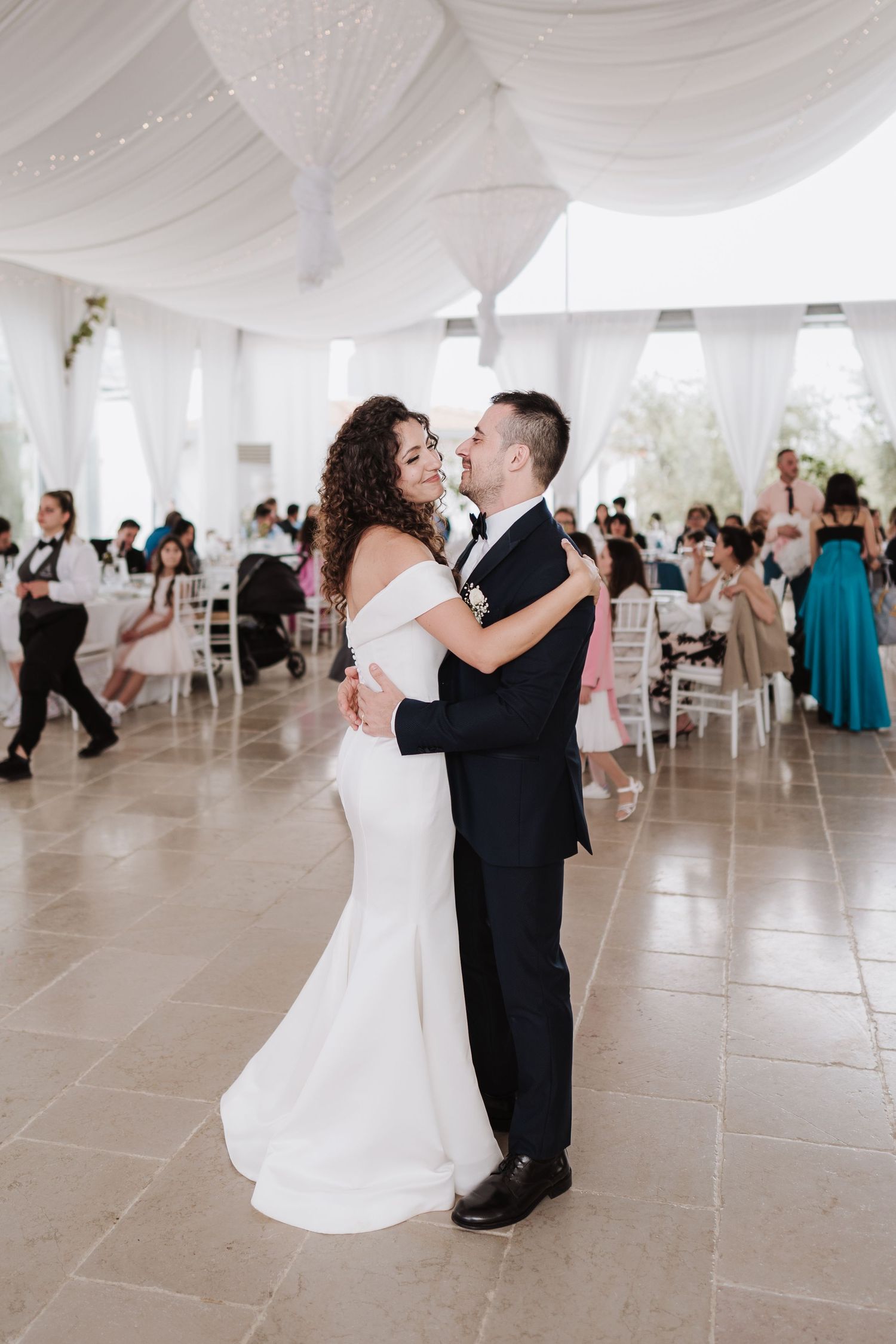 A couple embraces during their first dance in an elegant white-draped reception venue.