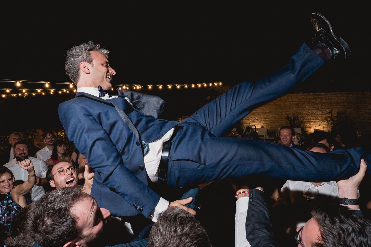 Guest in blue suit crowd surfing at nighttime party with string lights overhead.