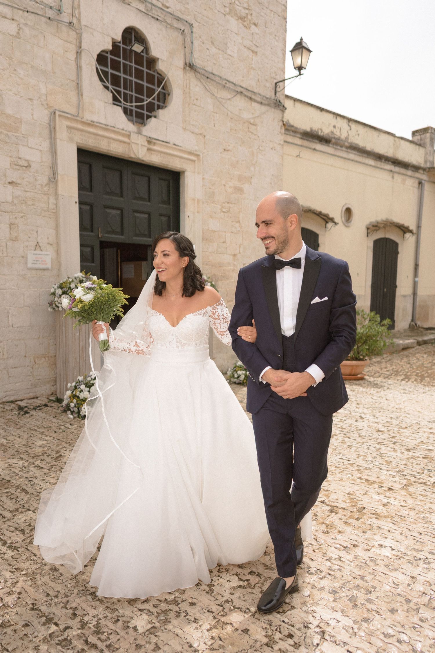 Newlyweds walking along cobblestone path outside historic stone building.