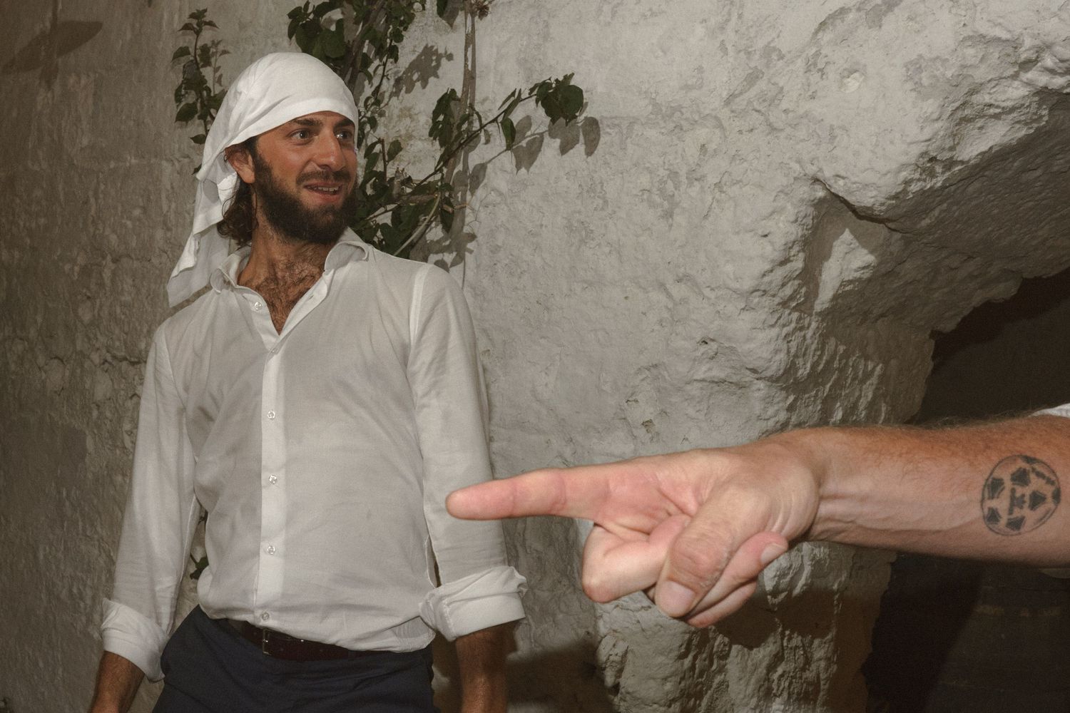A person in a white shirt and headwrap stands against a wall while a tattooed hand points in their direction.