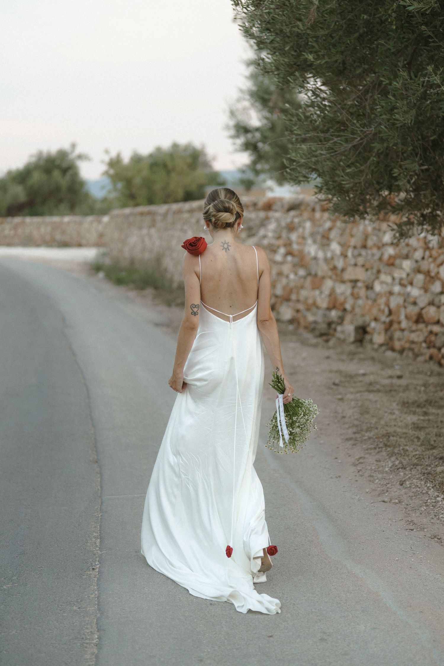 Bride in flowing white dress walking along countryside road holding bouquet.