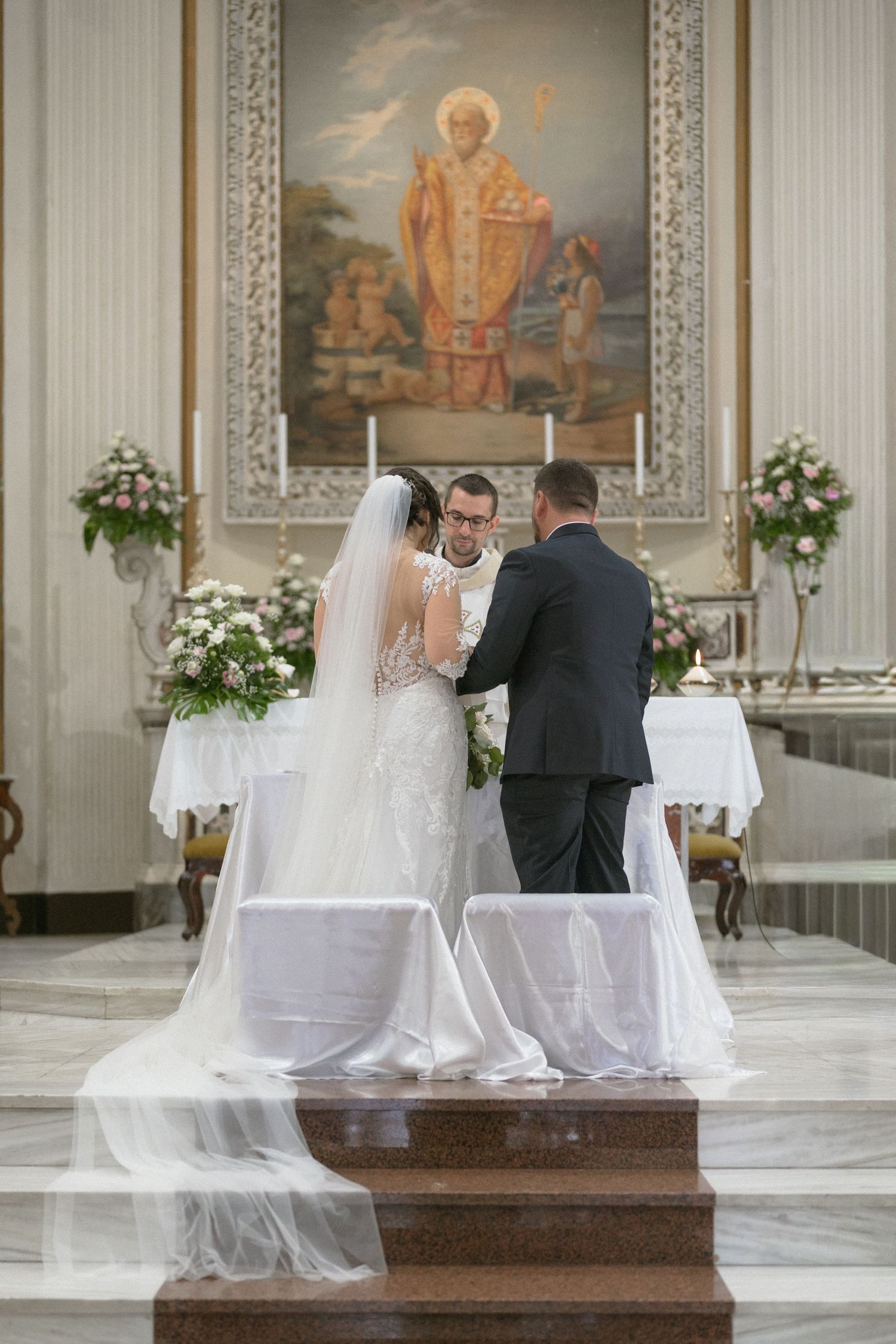 Wedding ceremony in an ornate church with religious paintings and white floral decorations.