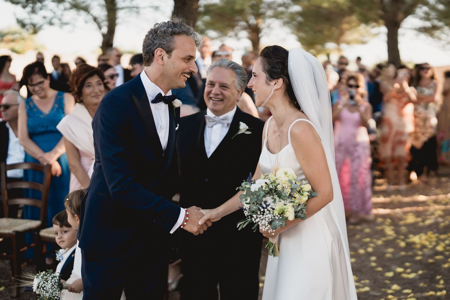 Outdoor wedding ceremony scene with couple exchanging vows under trees as guests look on.