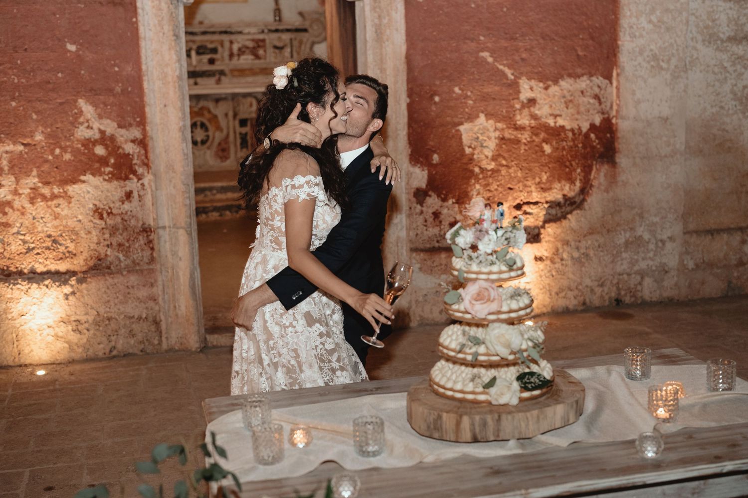 A wedding cake cutting scene with a tiered white cake decorated with flowers at a rustic venue.
