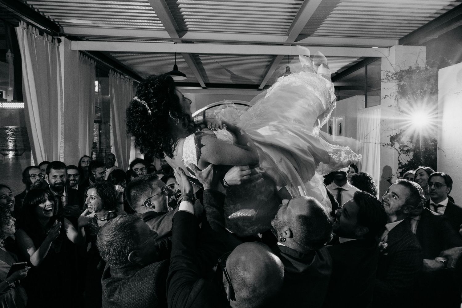 Groom lifts bride for a kiss surrounded by cheering wedding guests in black and white photo.