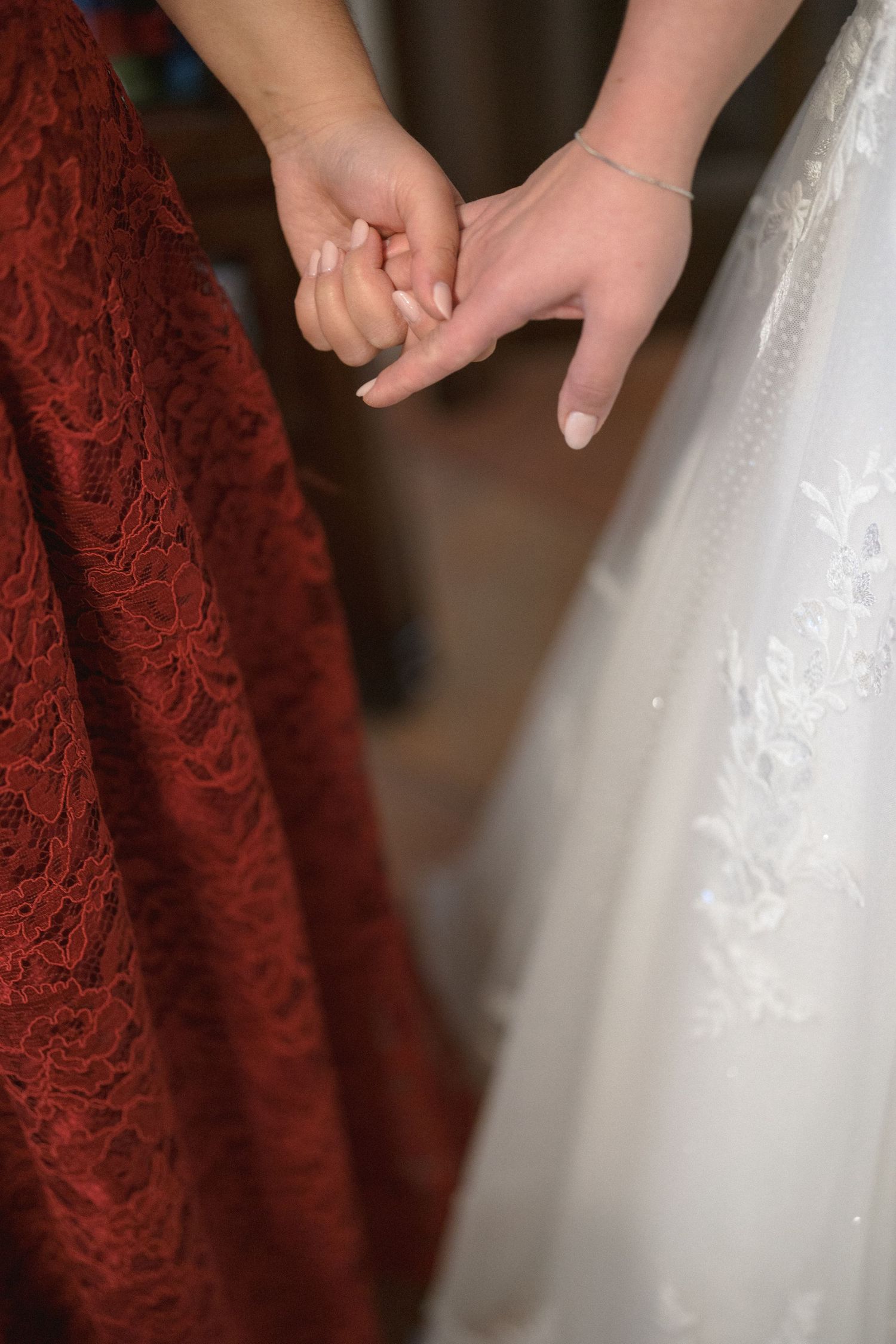 Bride in white lace gown holds hands with bridesmaid in red lace dress at wedding ceremony.