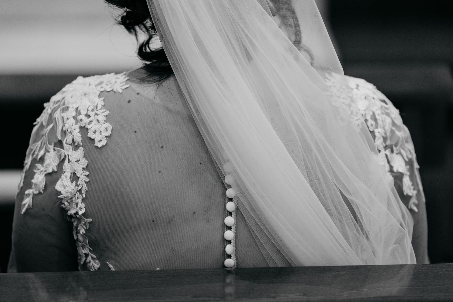 Black and white photo of a bride in a lace wedding dress with a flowing veil, viewed from behind.