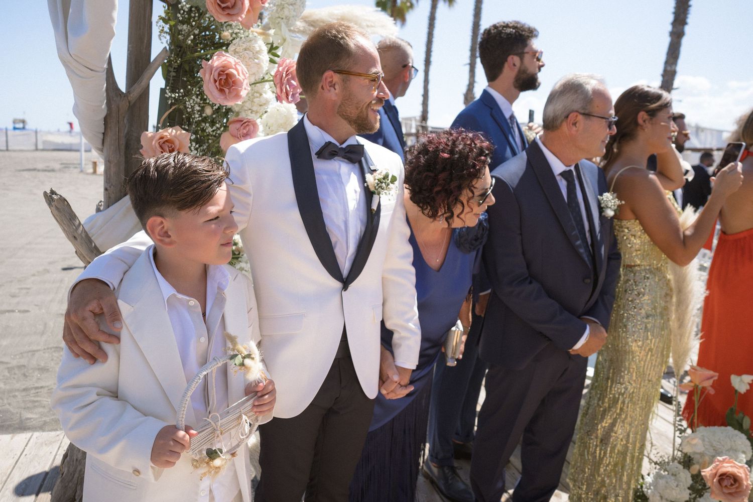 Wedding guests mingling outdoors with palm trees and floral decorations visible.