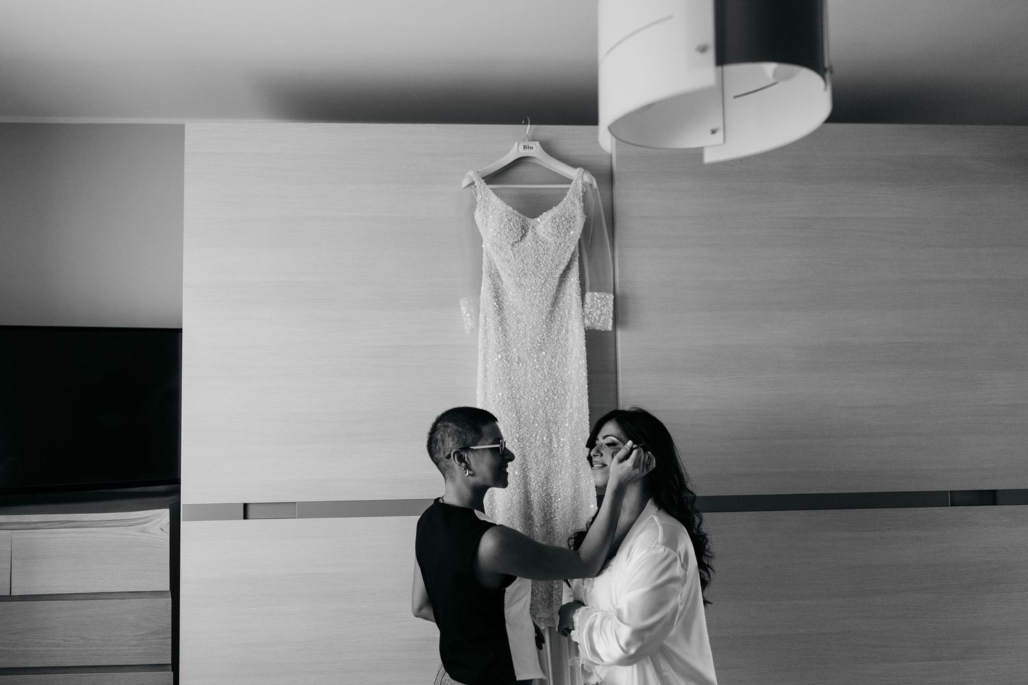 A black and white photo showing a couple preparing for their wedding with a dress hanging above.