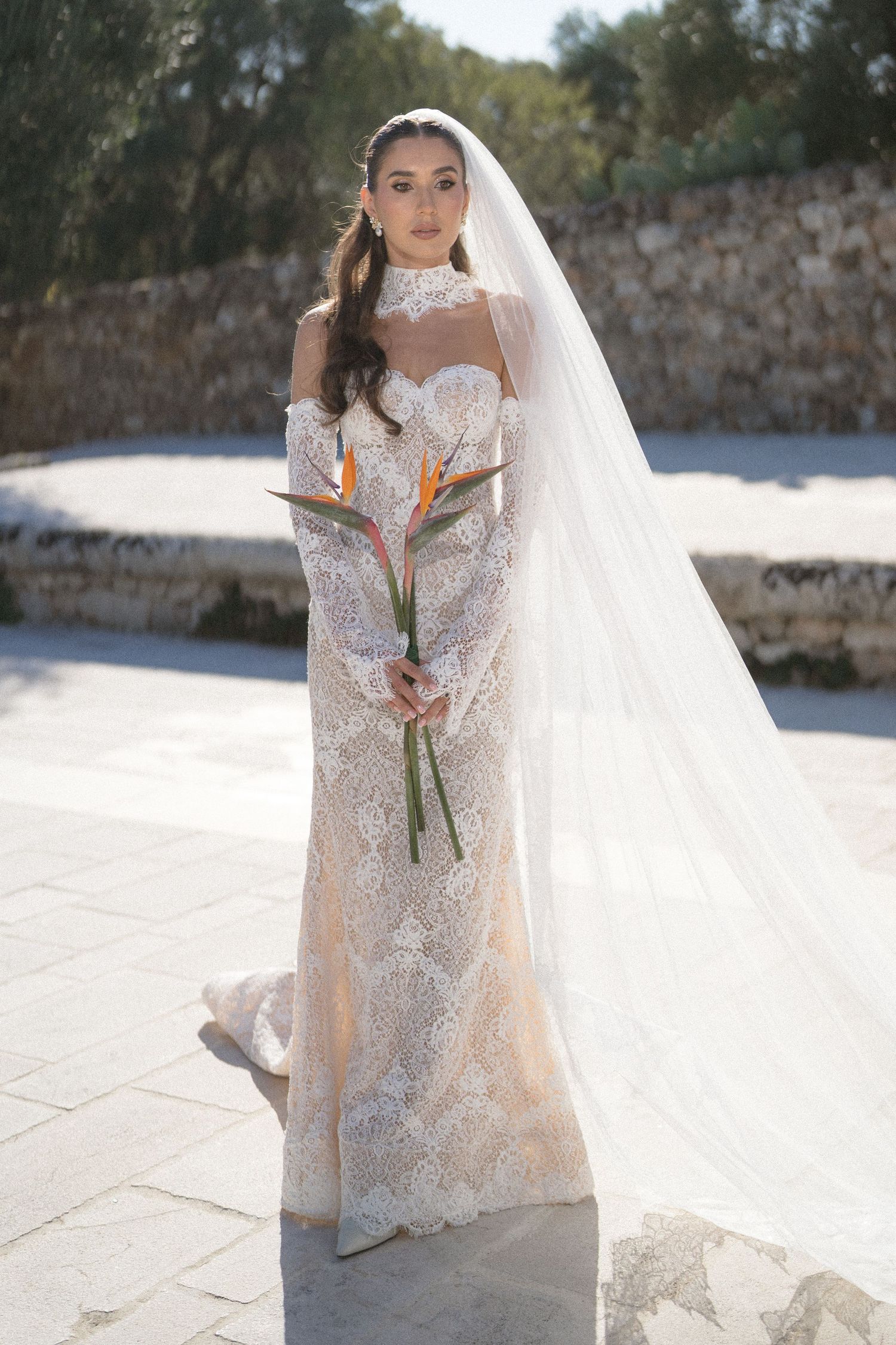 Bride in a vintage-style lace gown with high collar and long veil holds orange bird of paradise flowers on a sunlit stone terrace.