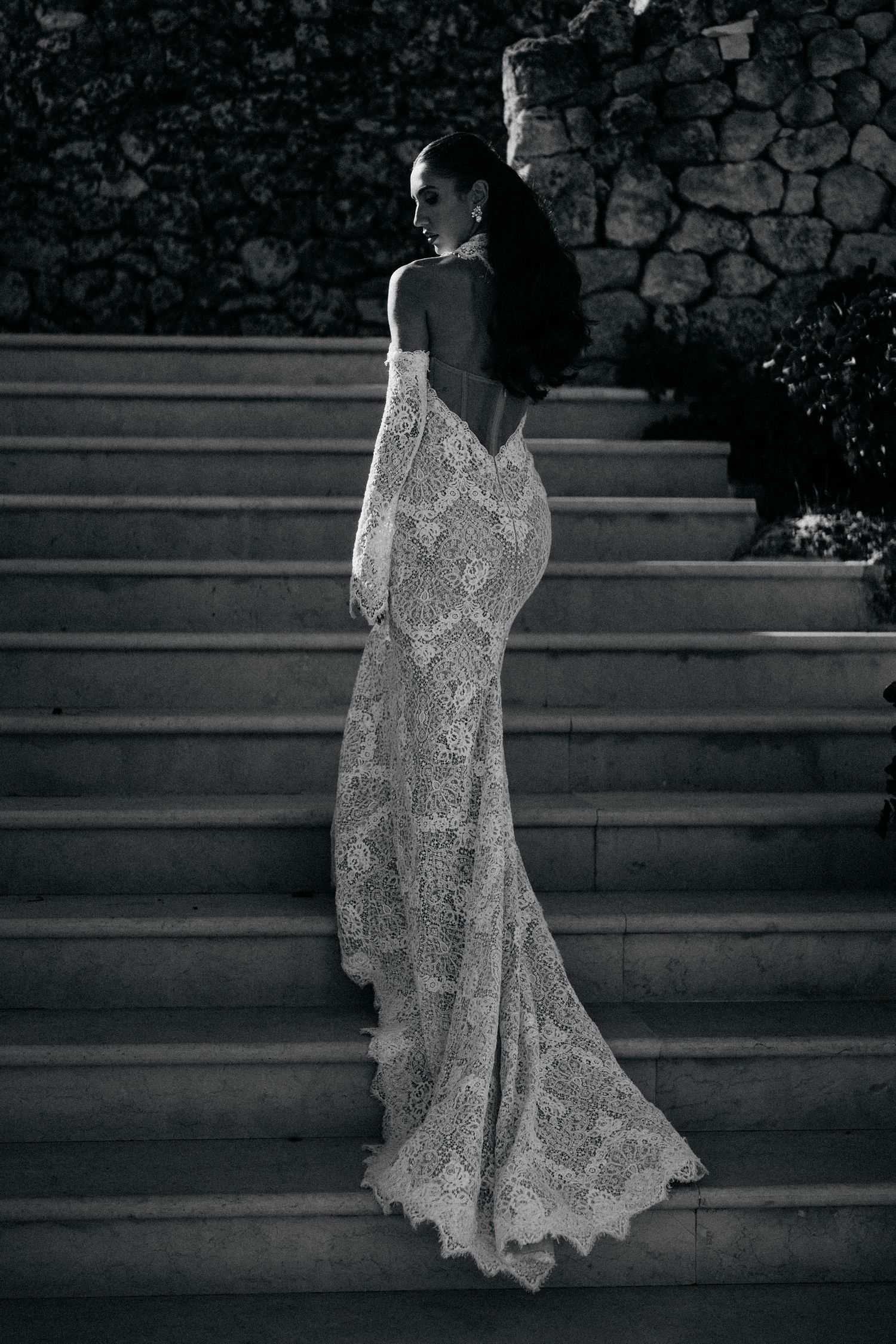 Black and white photo of a bride in a backless lace mermaid wedding dress with a long train, climbing stone steps outdoors.