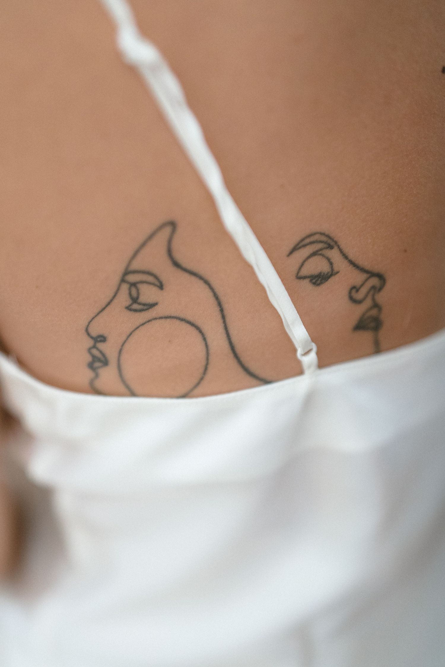 Close-up of minimalist line art face tattoos on a woman's back, partially covered by white dress straps.