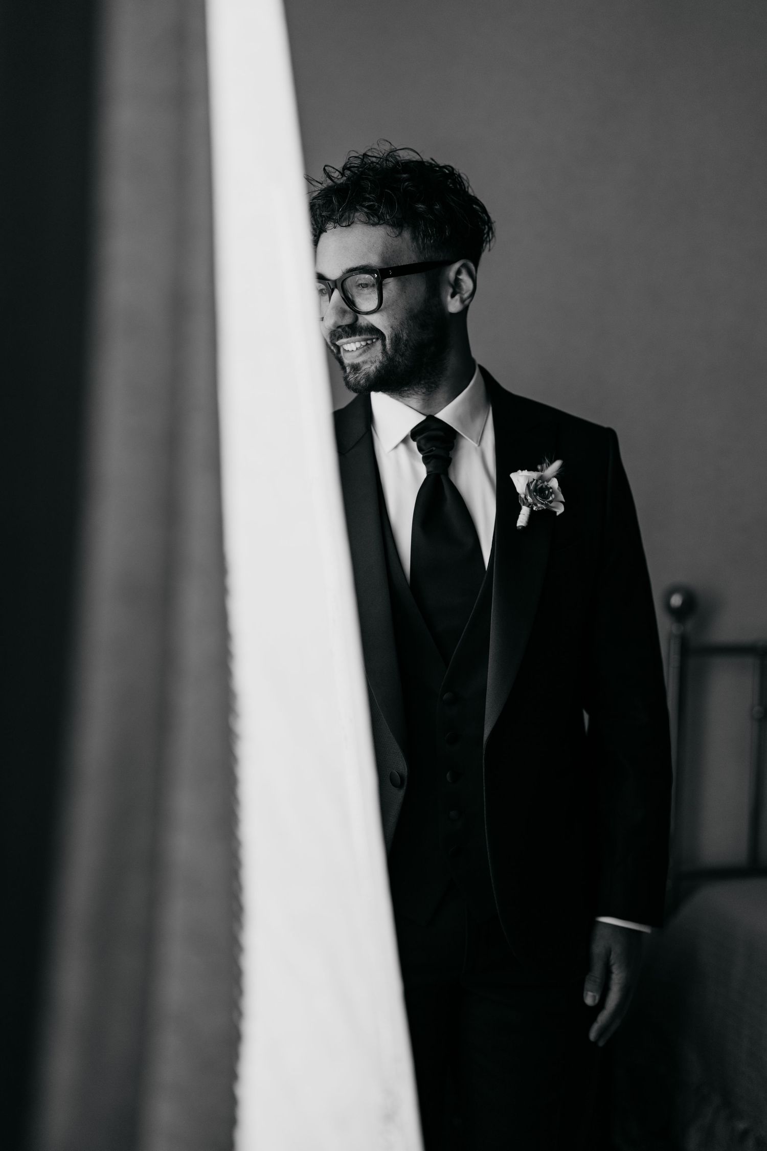 Smiling groom in black suit with boutonniere and glasses poses beside white drape in black and white wedding photo.