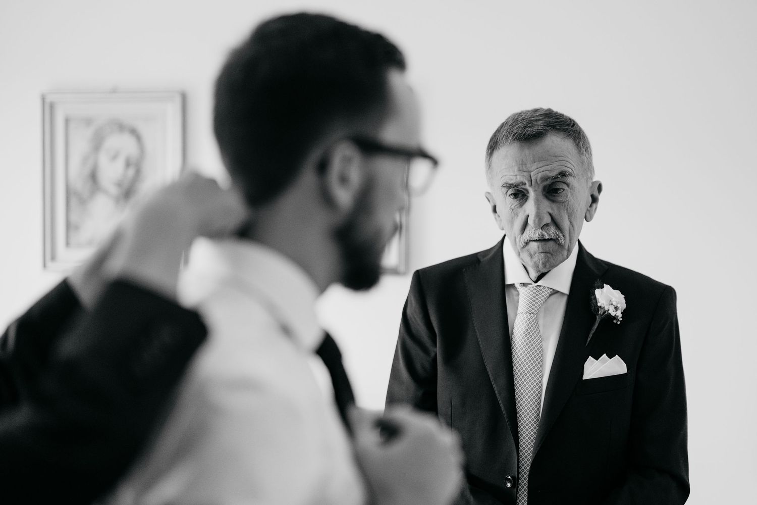 Black and white photo of two men in suits at a wedding, one adjusting his tie while an older man watches.