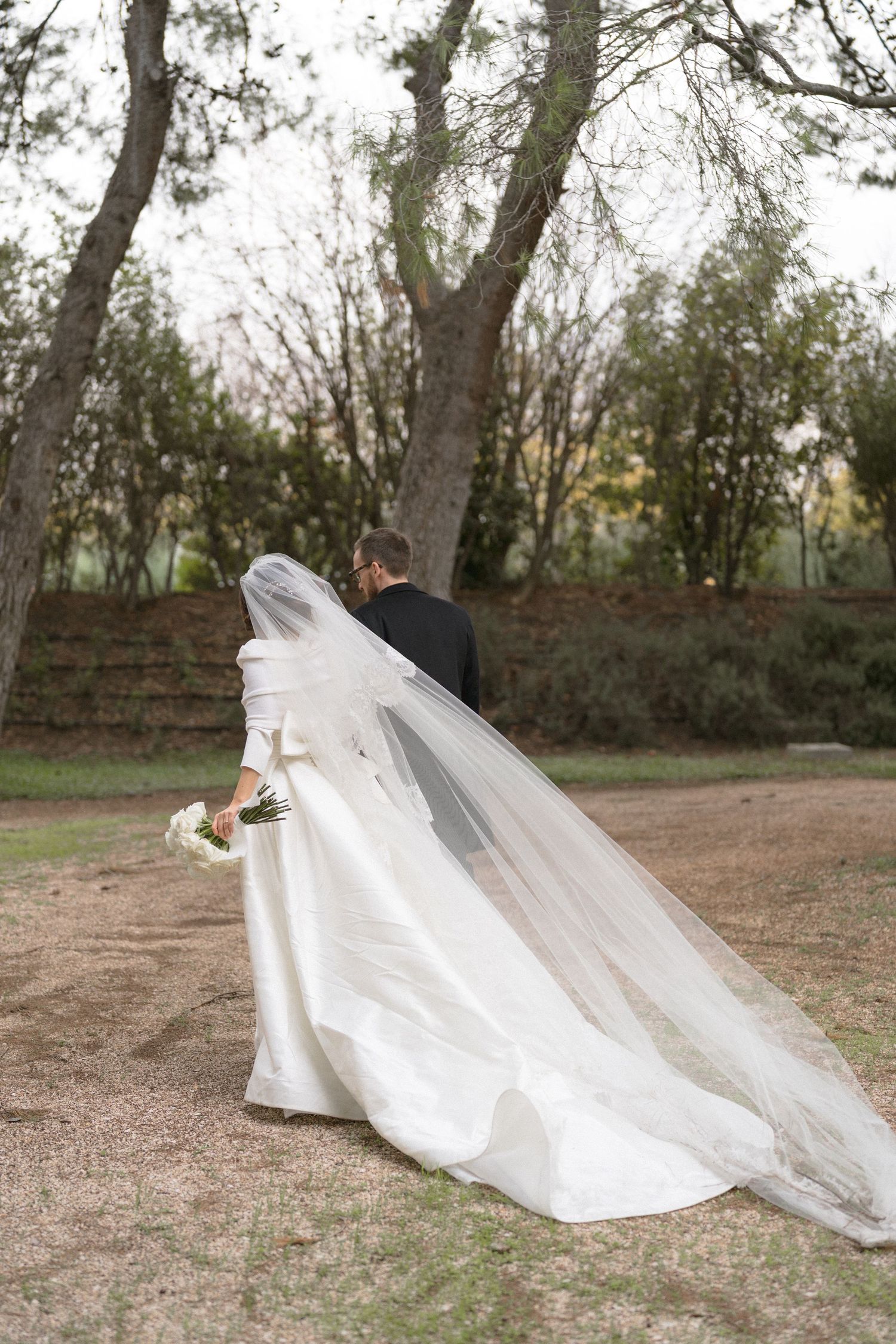 Bride and groom walking away in a garden, bride wearing a long white gown with flowing veil and bouquet.