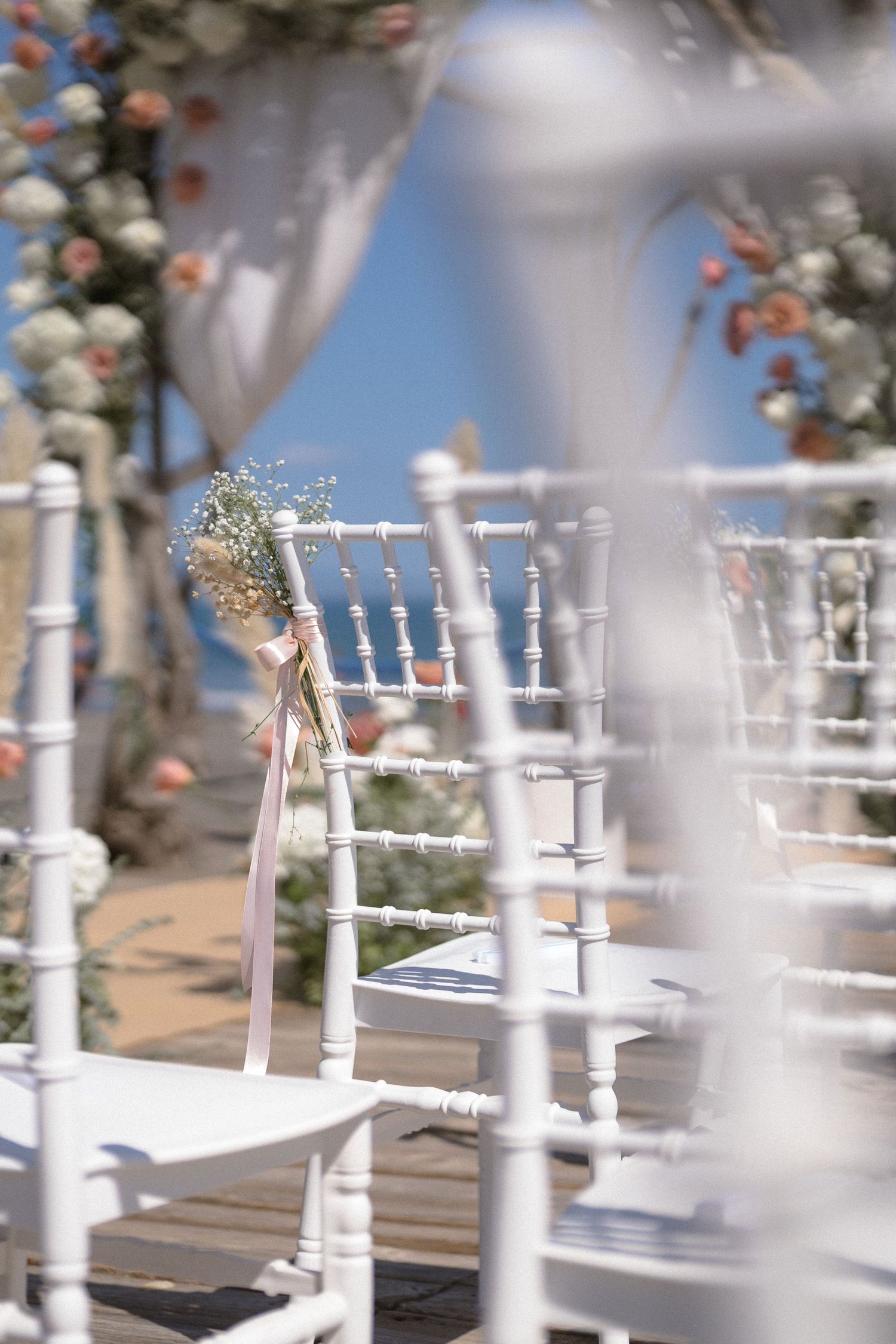 White chiavari chairs decorated with flowers set up for an outdoor wedding ceremony.