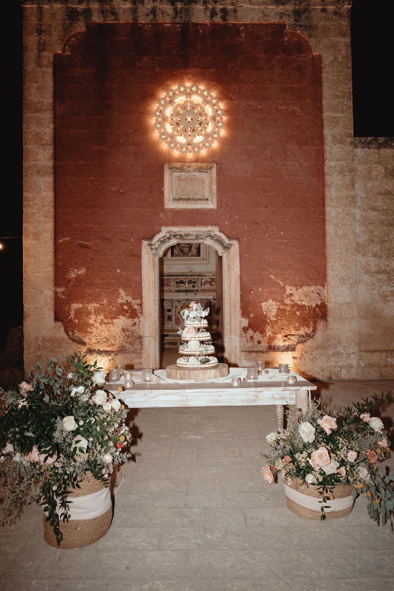 Wedding cake display setup against rustic brick wall with floral arrangements and chandelier lighting.
