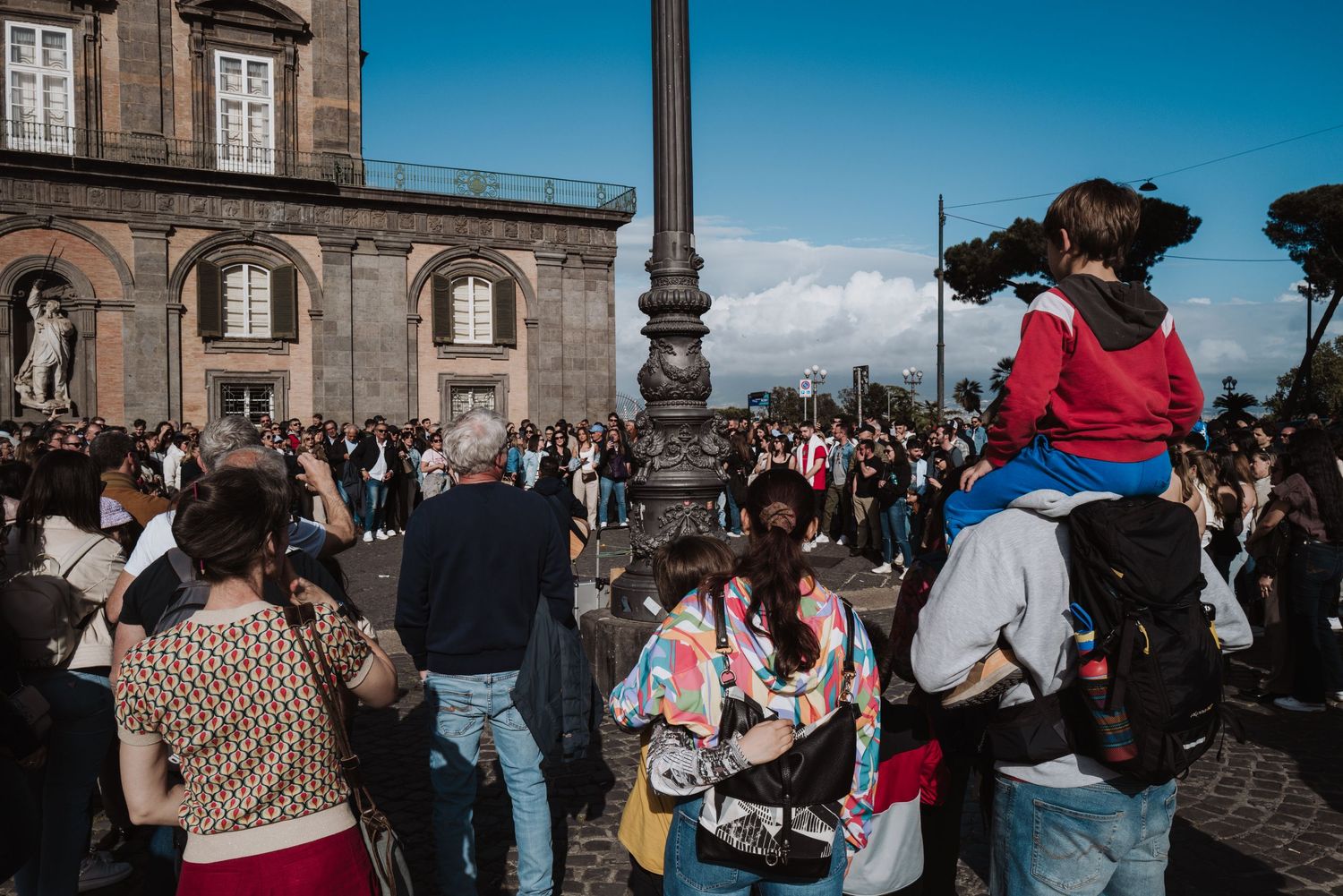 Tourists gather at a scenic viewpoint in Naples with Mount Vesuvius visible in the background on a sunny day.