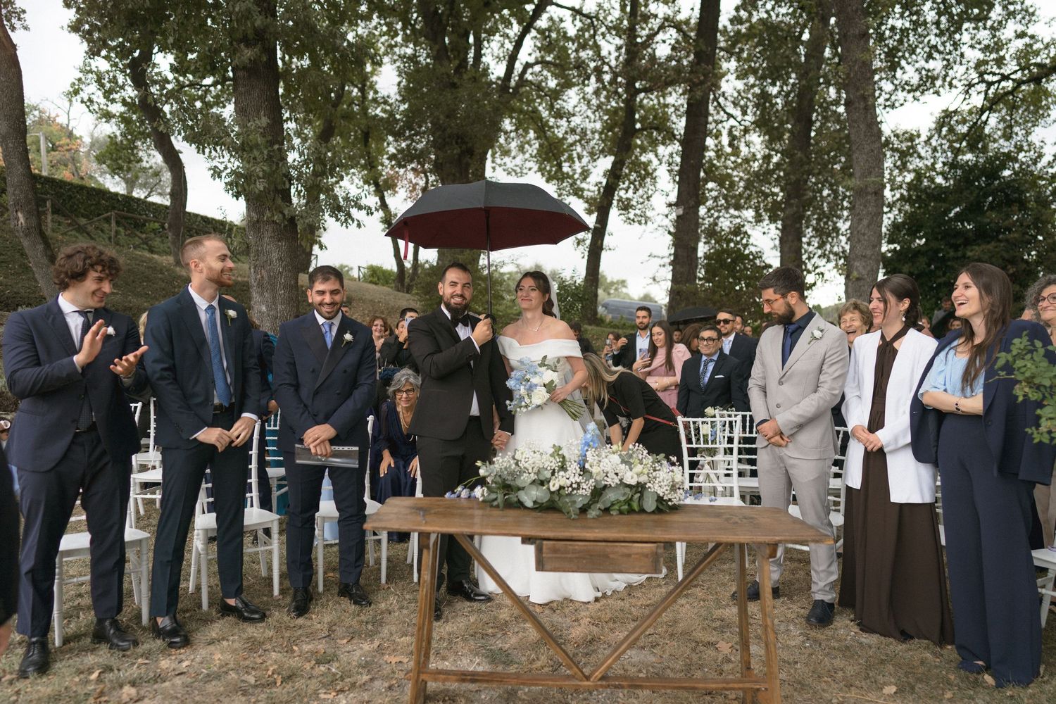 Wedding guests in formal attire standing in a line during an outdoor ceremony in a wooded setting.