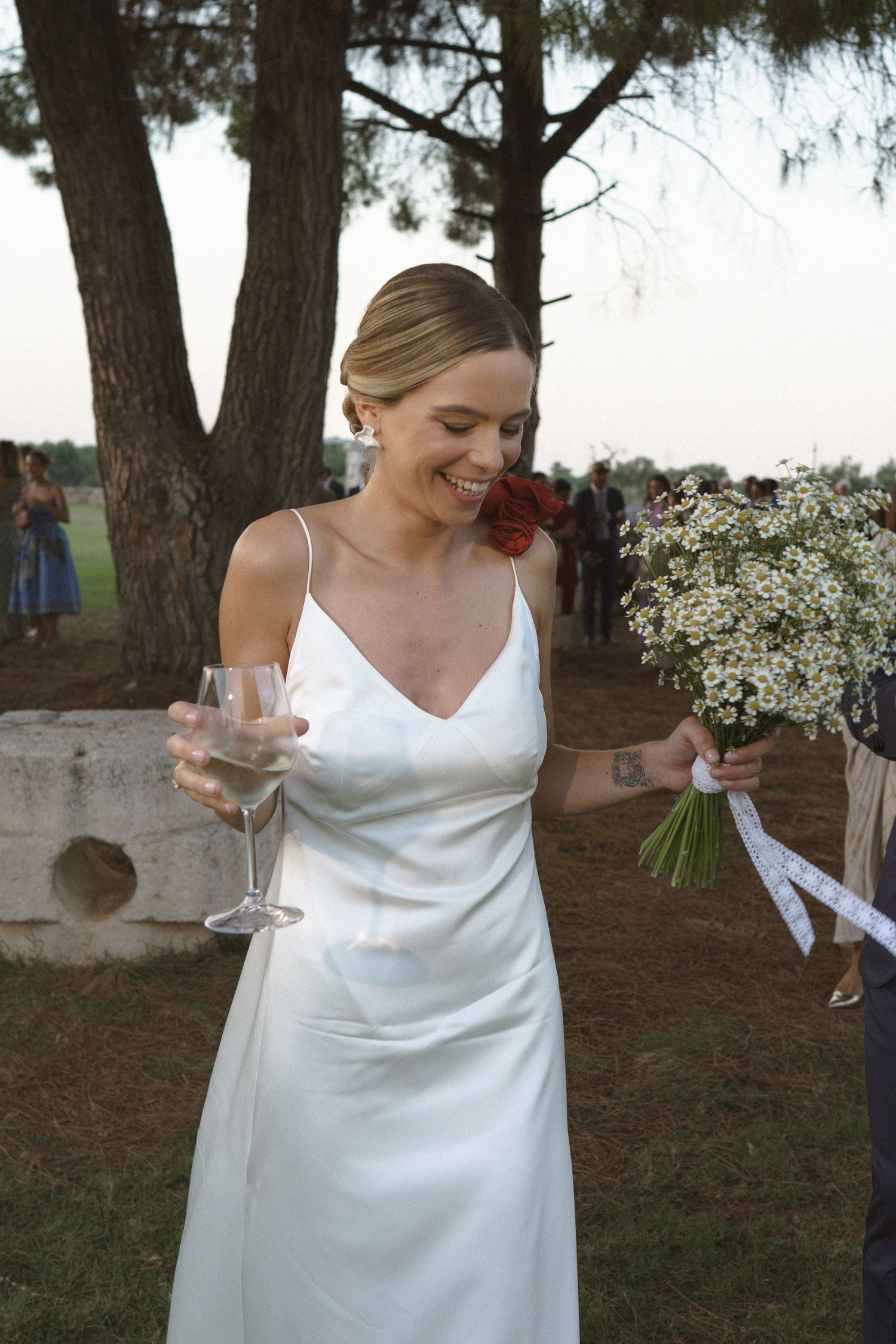 Woman in white silk wedding dress holds champagne glass and bouquet at outdoor garden celebration.