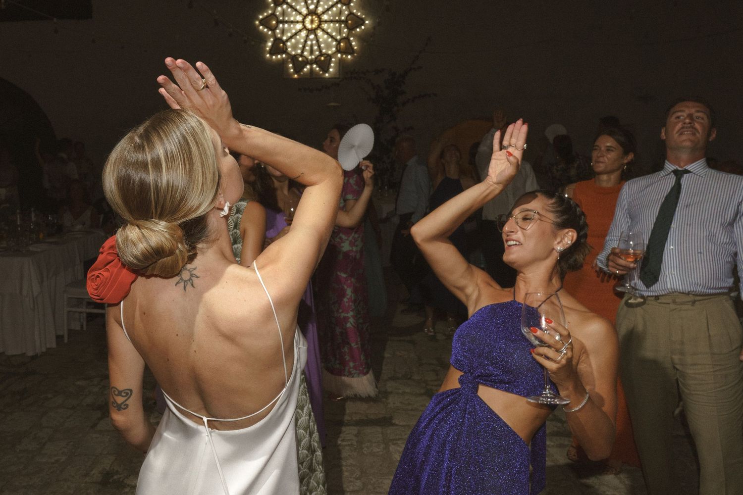 Wedding reception guests dance enthusiastically under decorative lighting at an evening celebration.
