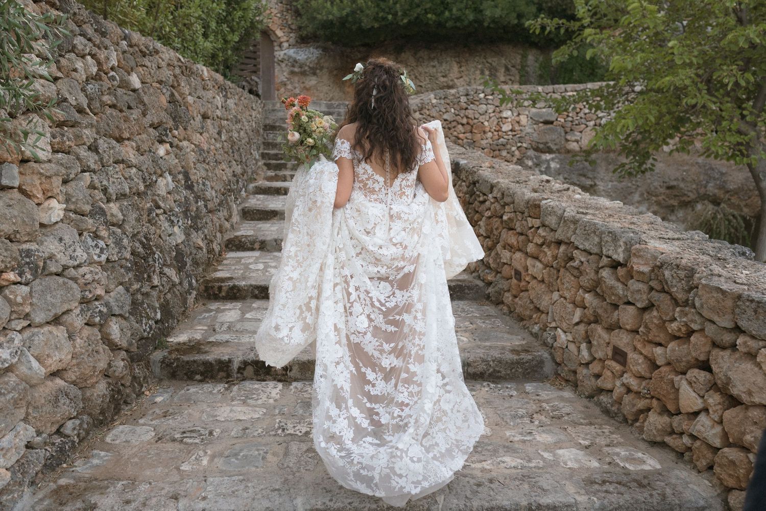 White lace wedding dress flowing down stone steps in Mediterranean setting.