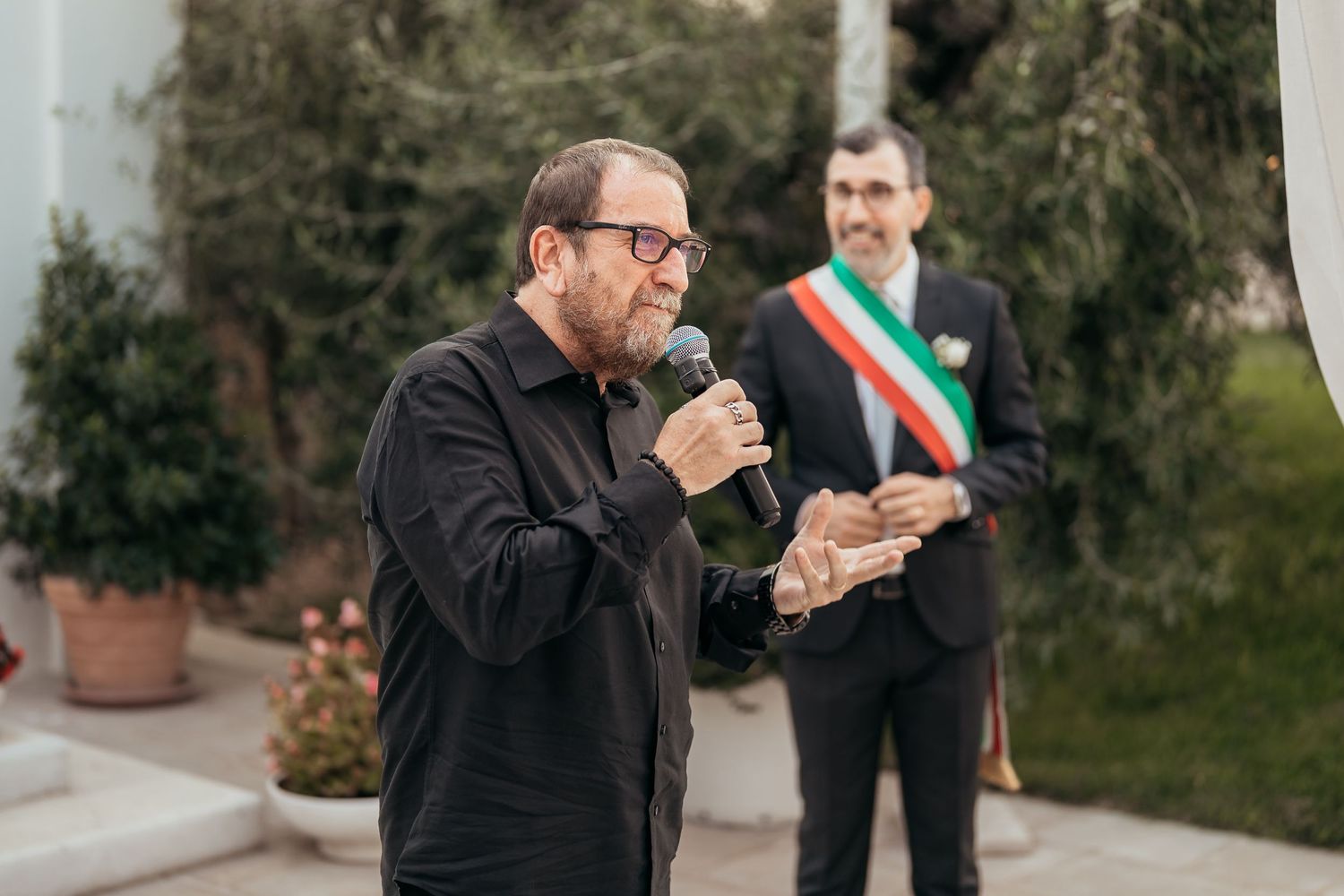 A speaker addresses the crowd during an outdoor ceremony while an official with a sash stands nearby.