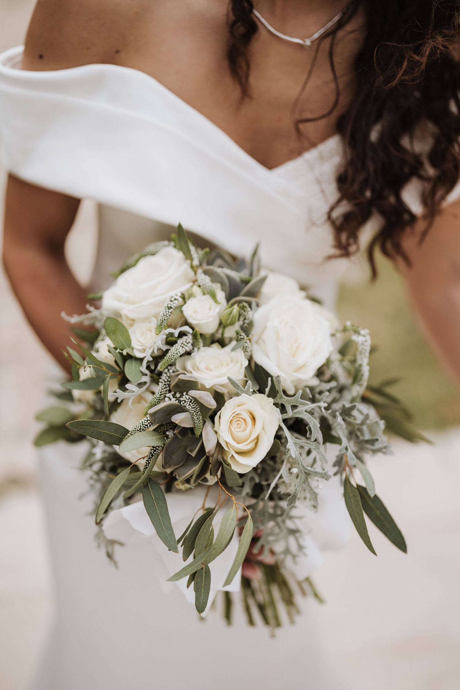 A delicate white rose wedding bouquet with eucalyptus and greenery against a white dress.