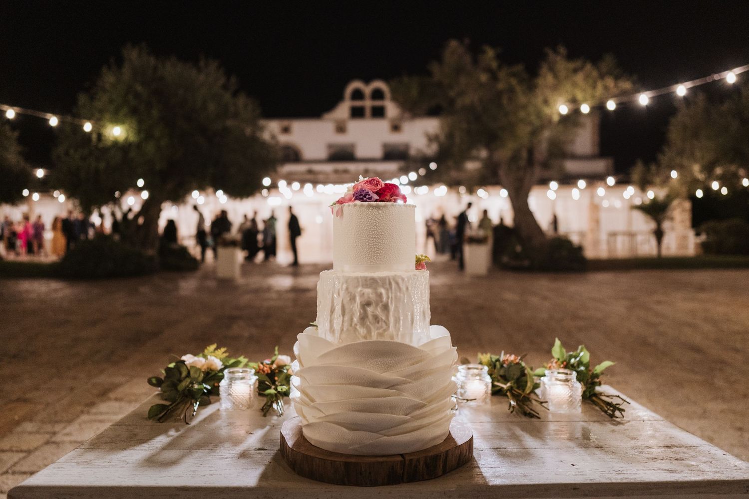 Elegant white wedding cake displayed outdoors at night with string lights and a venue in the background.
