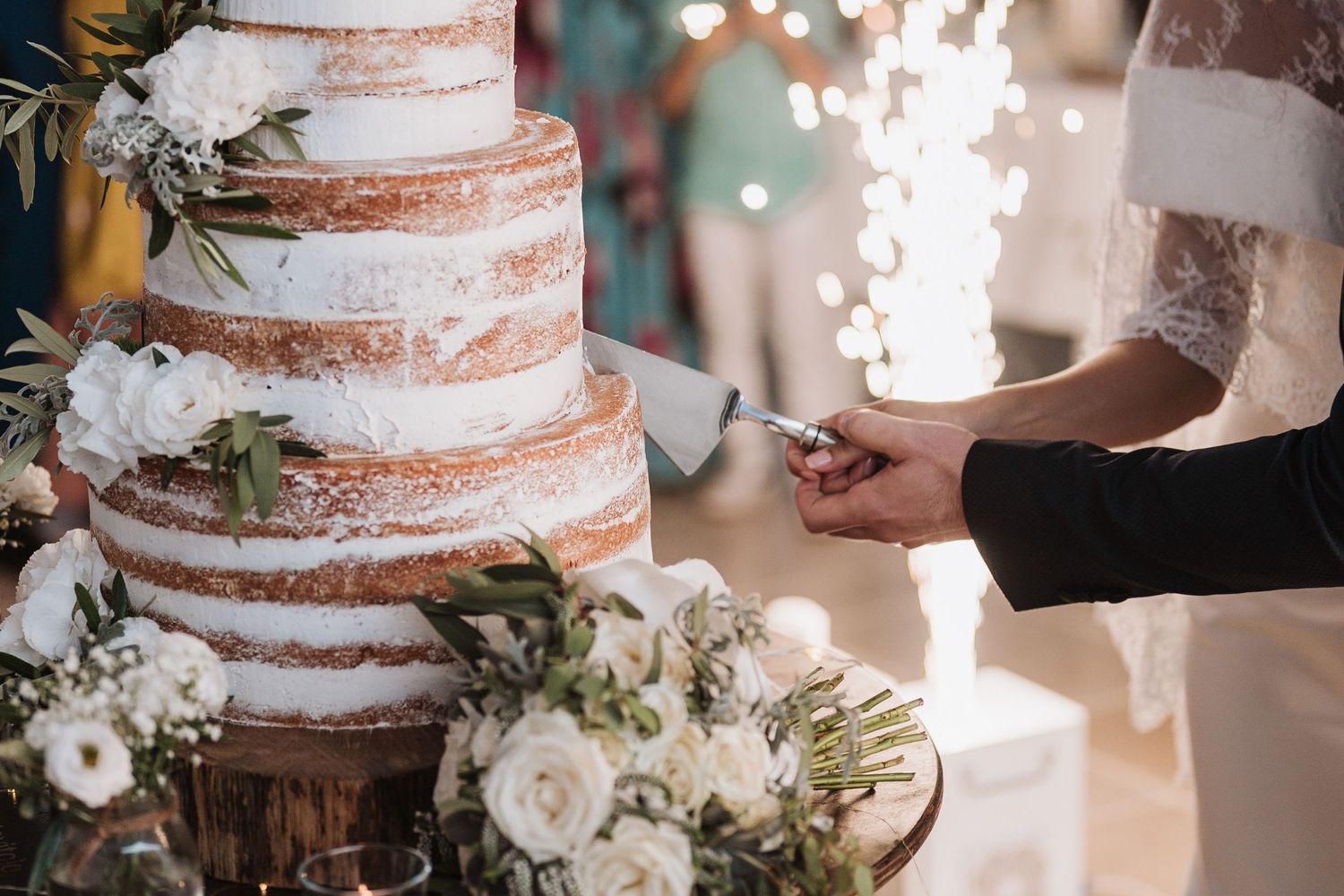 A rustic naked wedding cake decorated with white flowers and greenery being cut while sparklers glow in the background.
