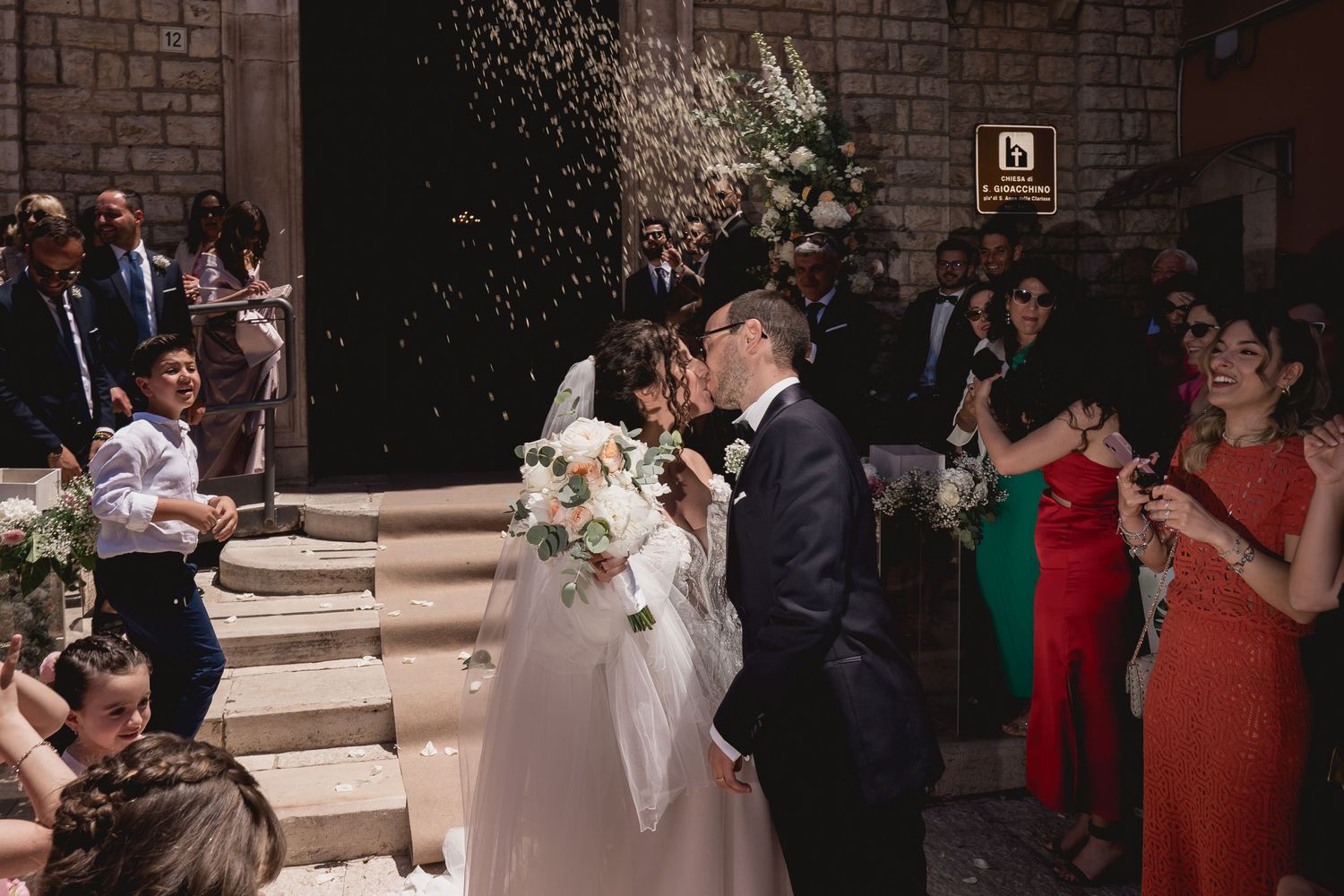 Guests celebrate as the newlyweds share their first kiss on church steps amid rice throwing and flowers.
