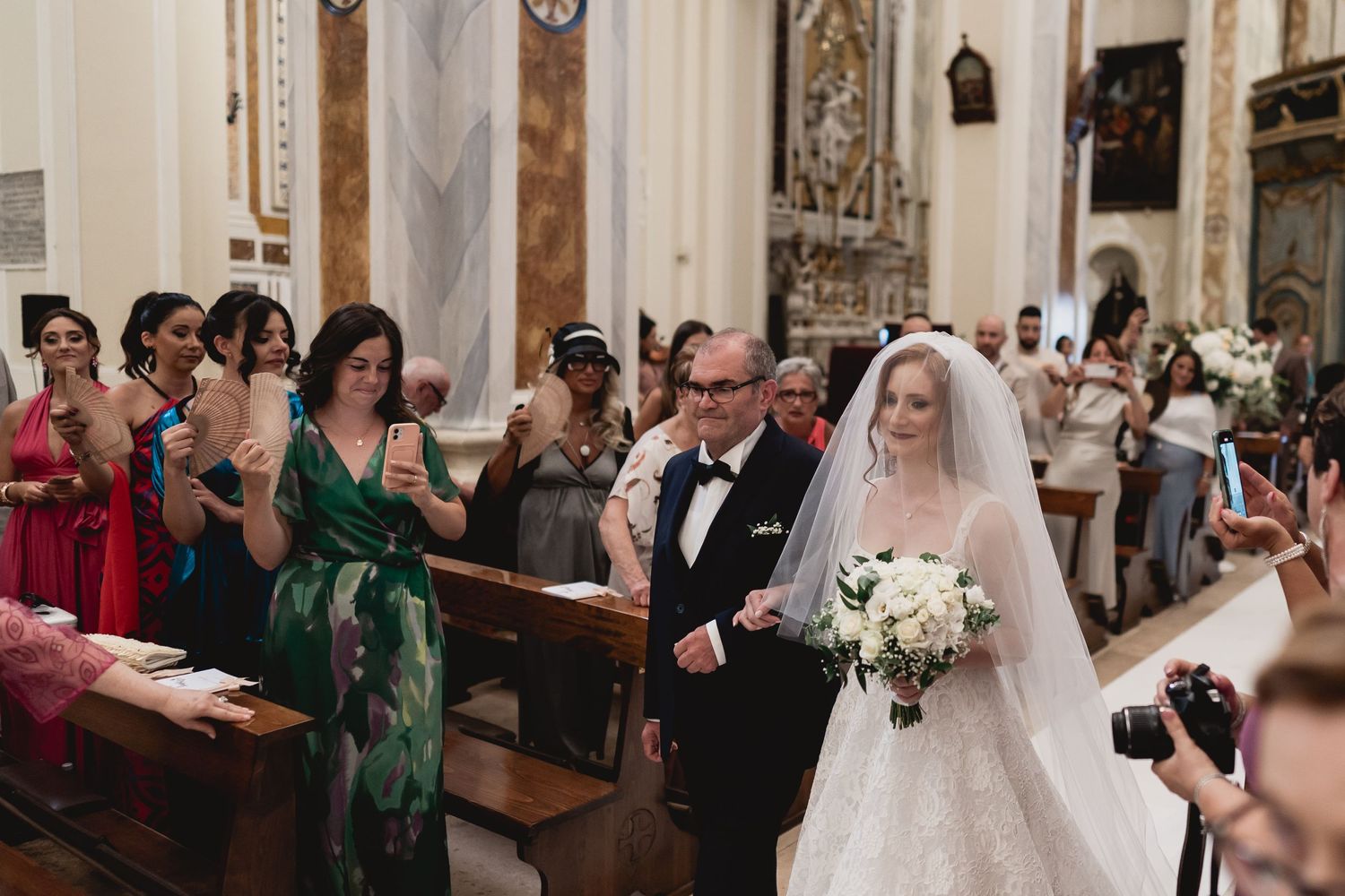 Wedding ceremony procession in ornate church with guests watching from wooden pews.