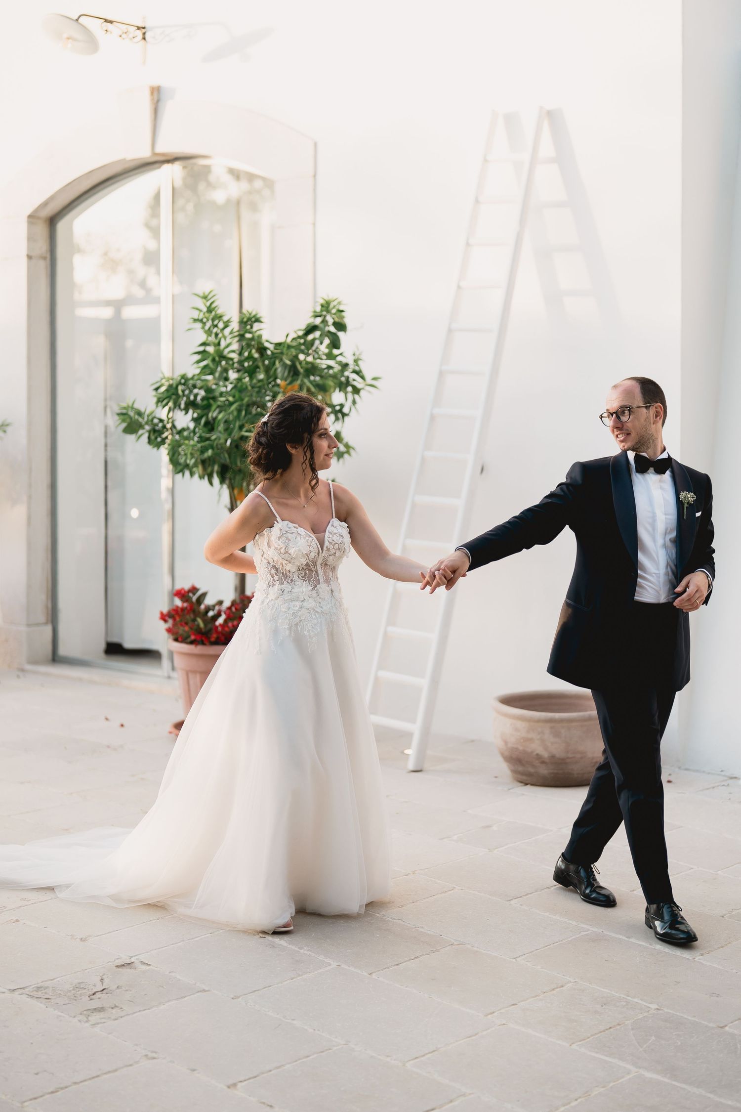A romantic wedding moment with a couple dancing in an elegant white venue with a ladder and potted plants visible.