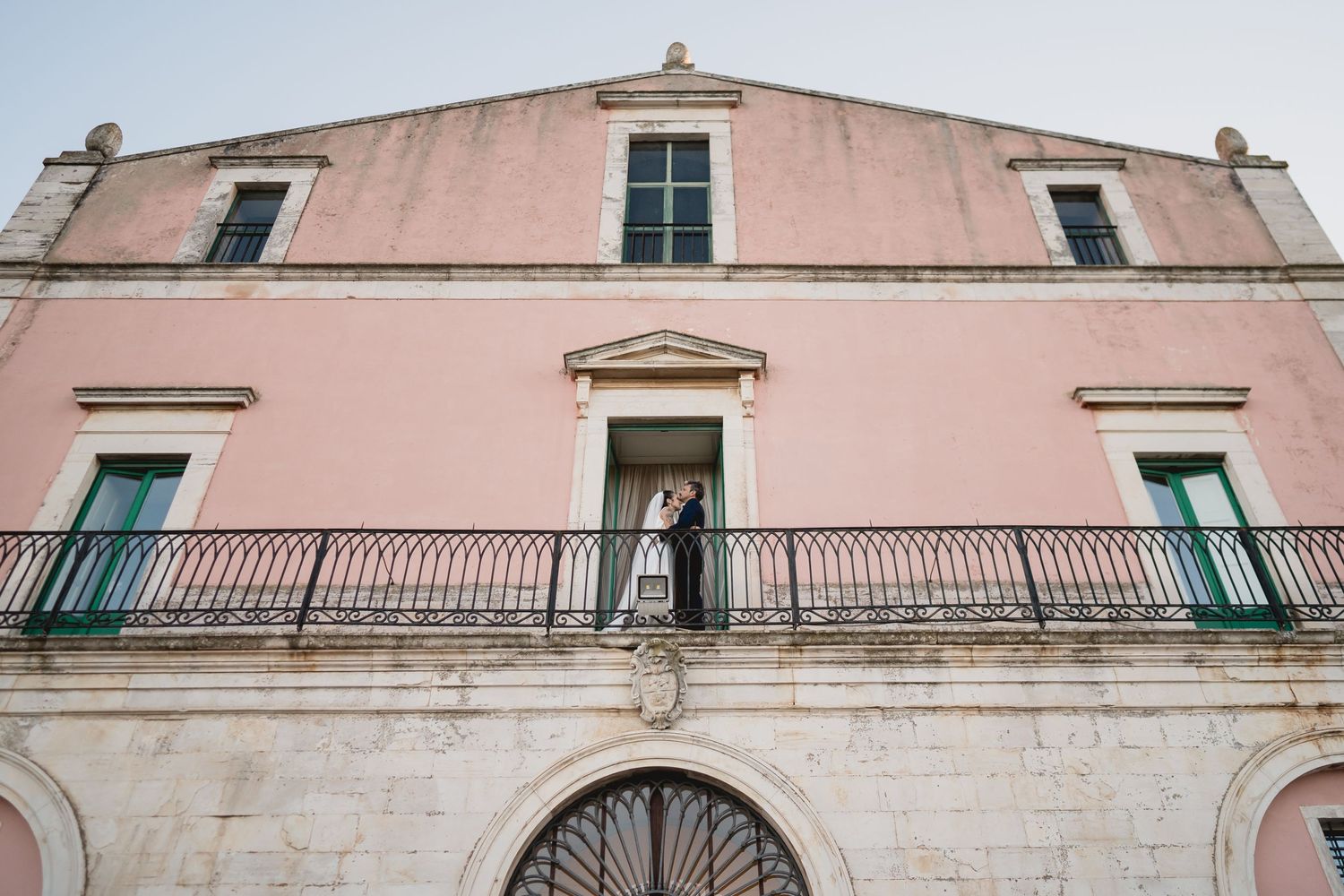 A figure stands on an ornate balcony of a vintage pink Mediterranean building with green shutters and arched doorway.