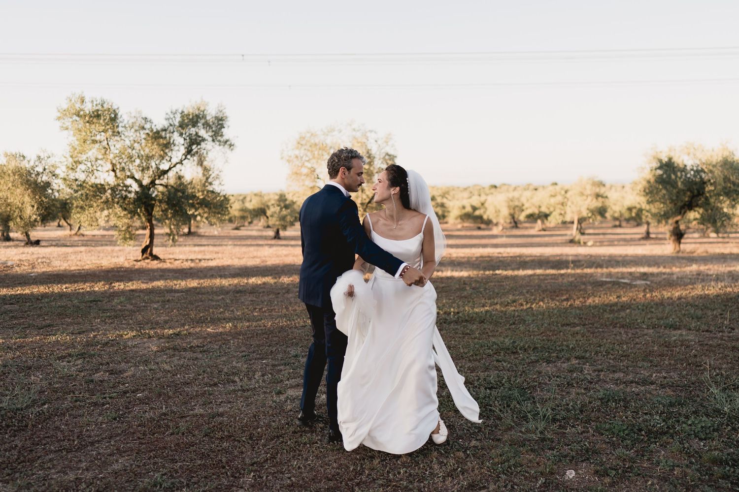 A couple poses together in an olive grove during golden hour.