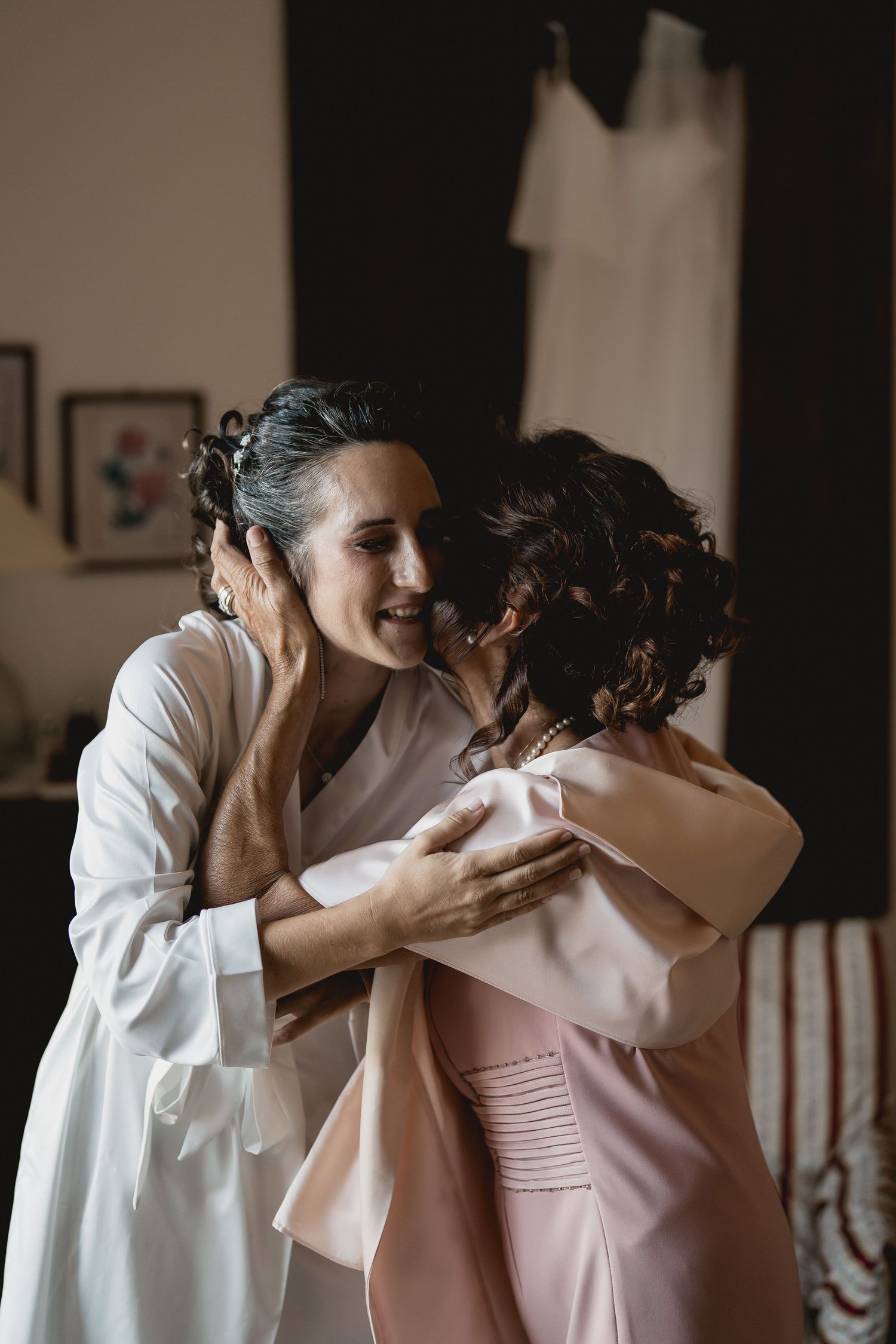 Two people wearing white and pink embrace warmly in a dimly lit room with a dress hanging in background.