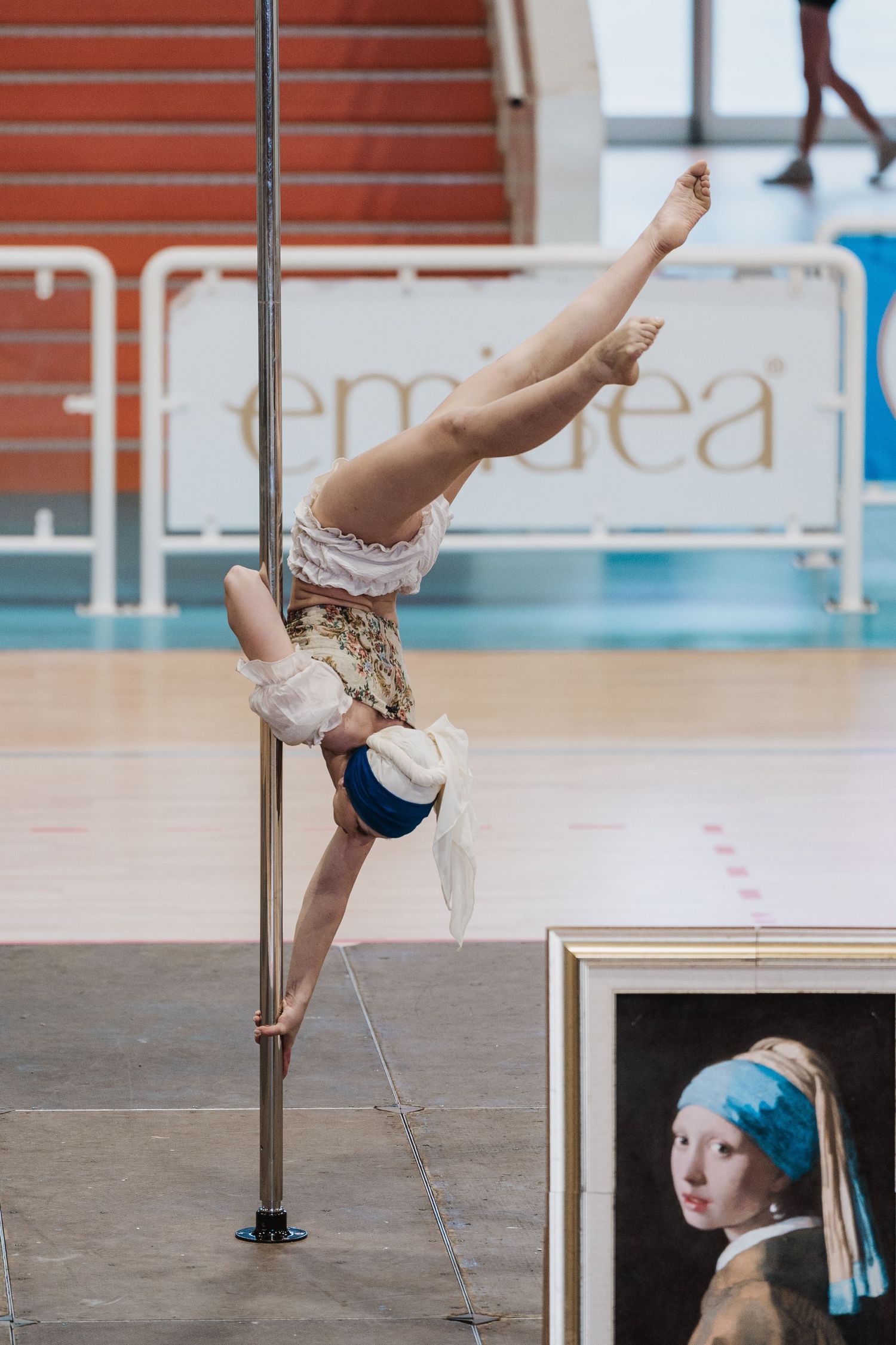 A pole dancer performs a graceful inverted split pose next to a framed Girl with Pearl Earring painting reproduction.