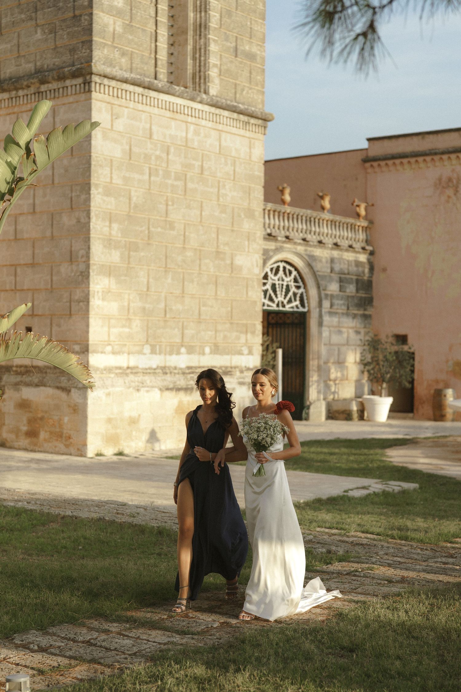 Two people in elegant dresses pose together on the grounds of a historic stone church with ornate architecture.