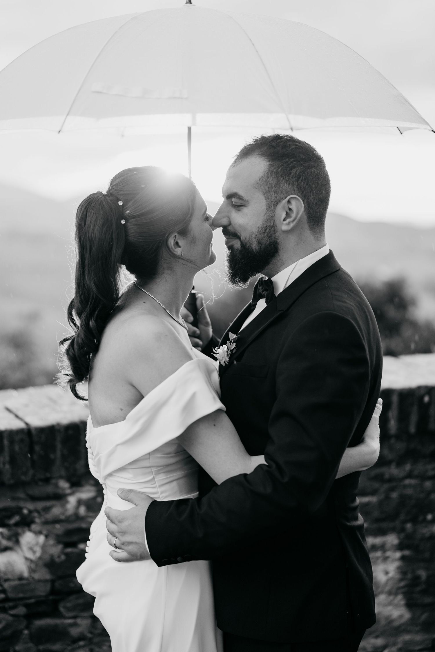 A black and white wedding photo captures an intimate moment between newlyweds under a white umbrella.