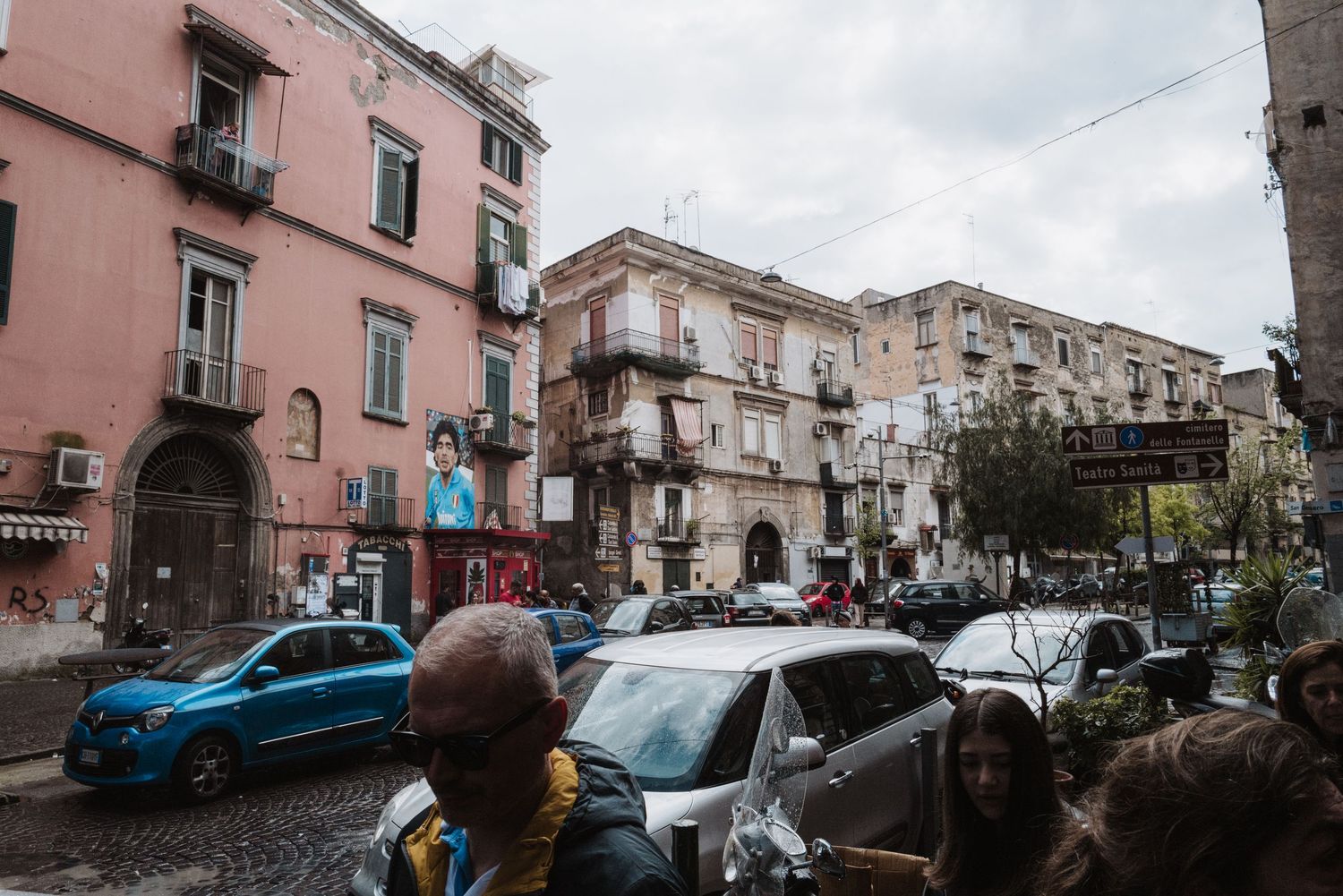 Busy street scene in Naples with pink buildings and parked cars under cloudy skies.