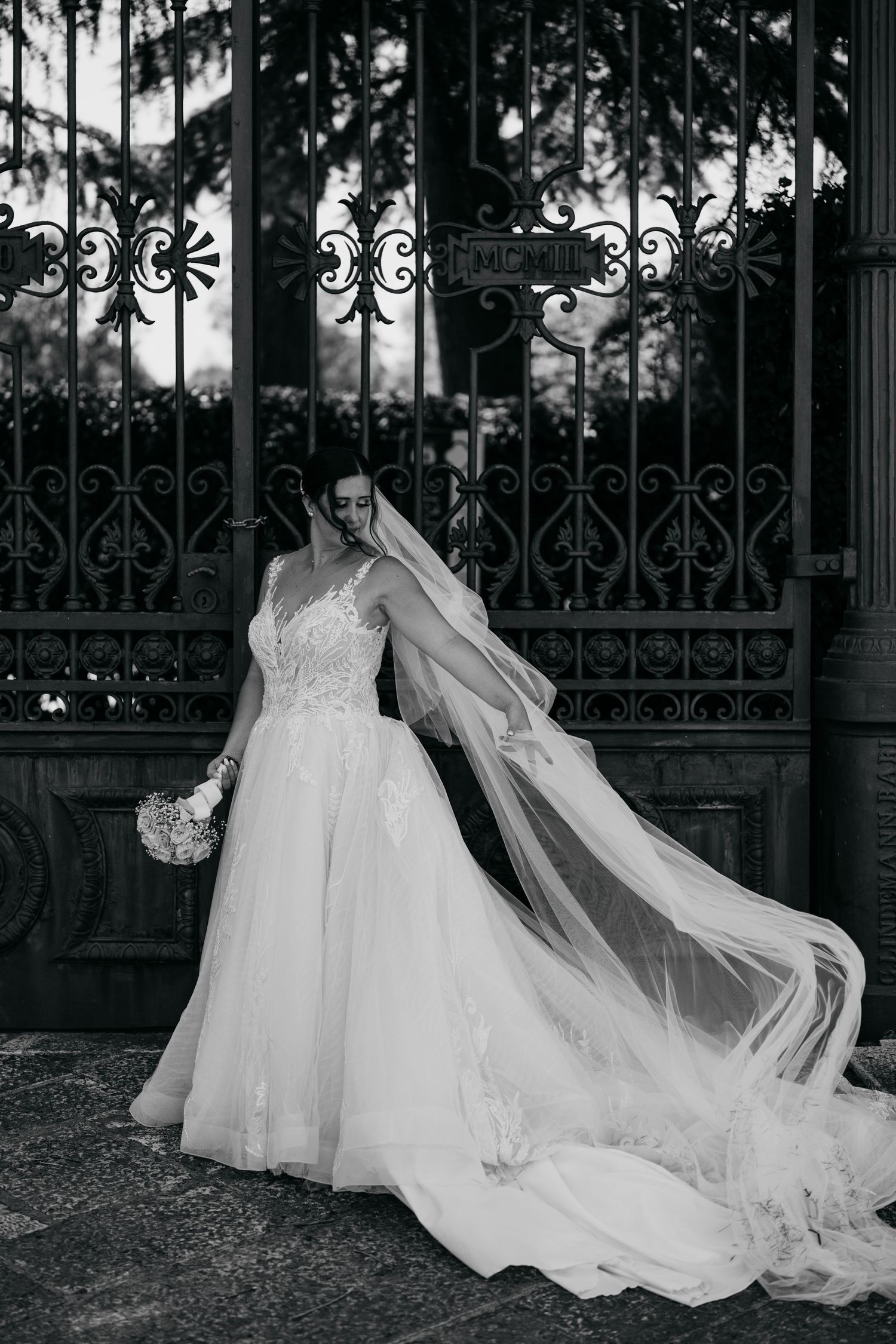 Black and white photo of a wedding dress with flowing veil against ornate iron gates.