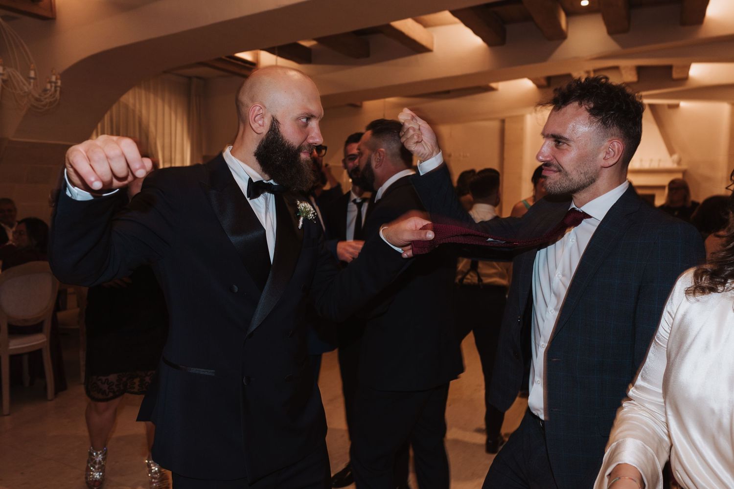 Wedding guests in suits share a playful moment during a reception celebration indoors.