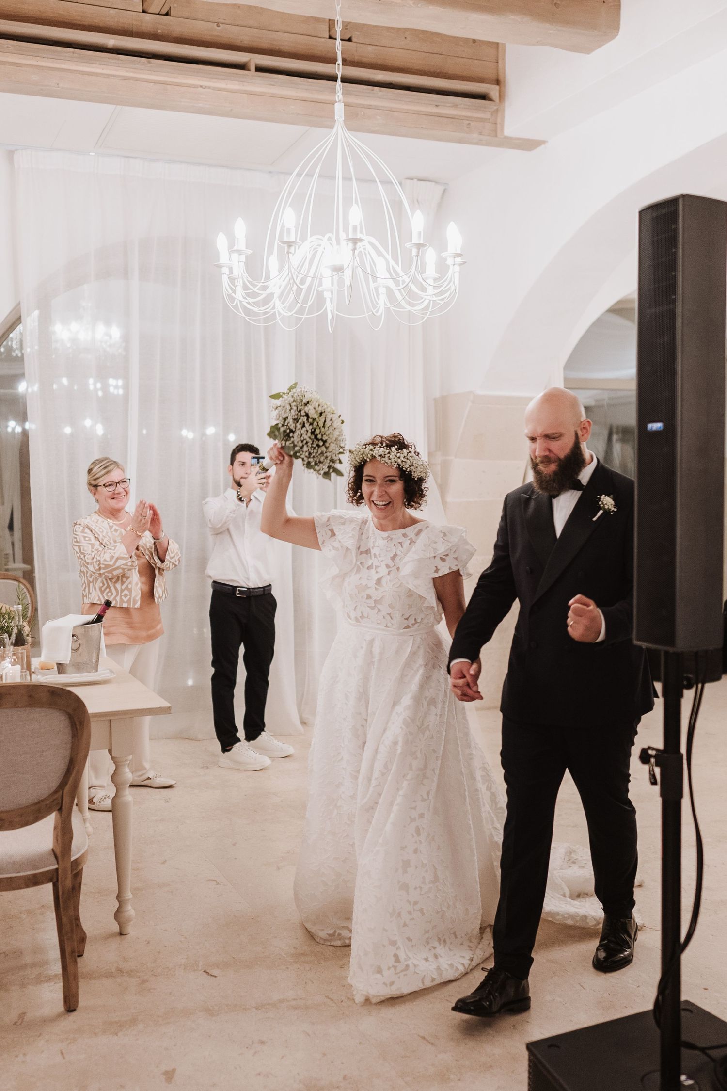 Wedding celebration in an elegant white room with chandelier and wooden beam ceiling.