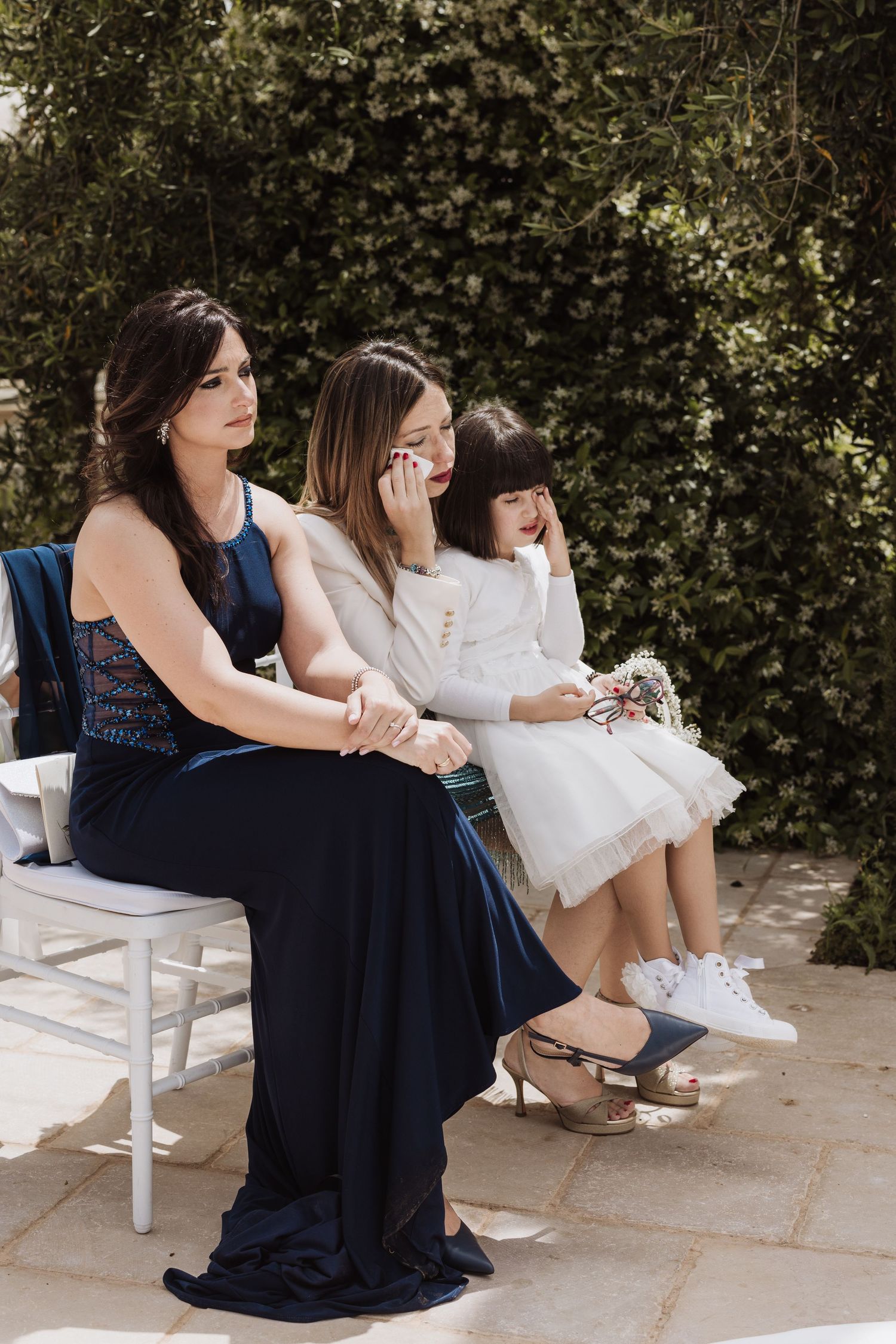 Wedding guests in formal attire seated on white chairs during outdoor ceremony.